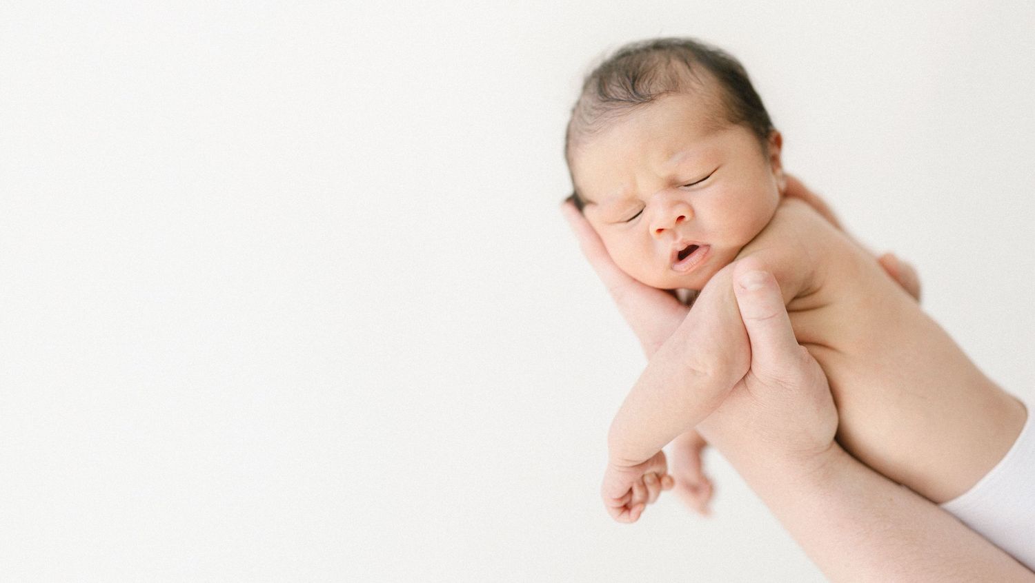 Newborn baby sleeping peacefully while being gently held in adult hands against white background.
