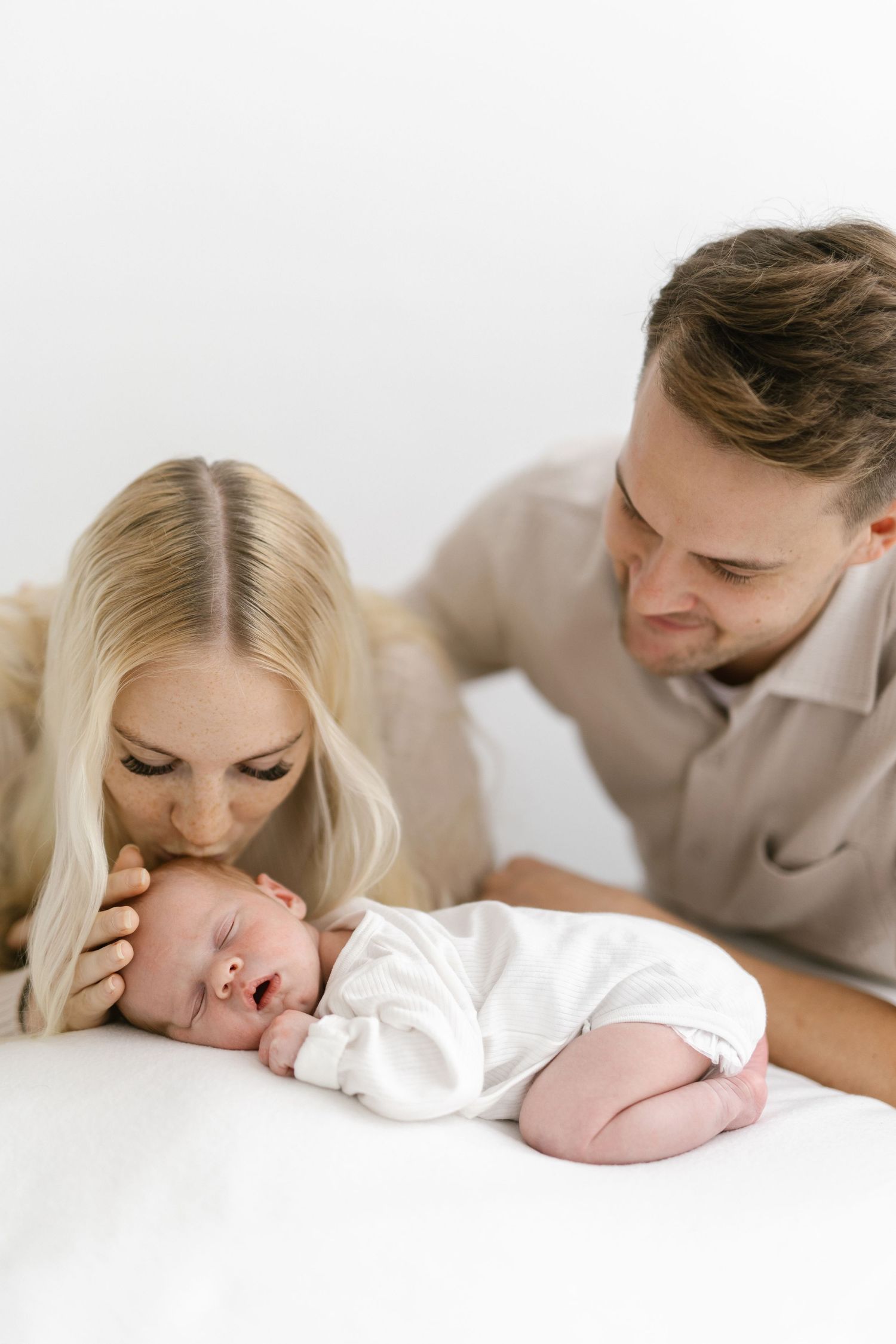 A young couple lovingly gazes at their sleeping newborn baby dressed in white on a soft surface.