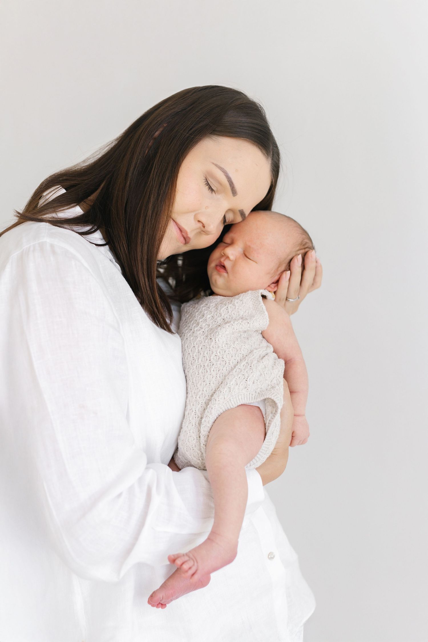 Mother in white embracing sleeping infant against minimalist white background.