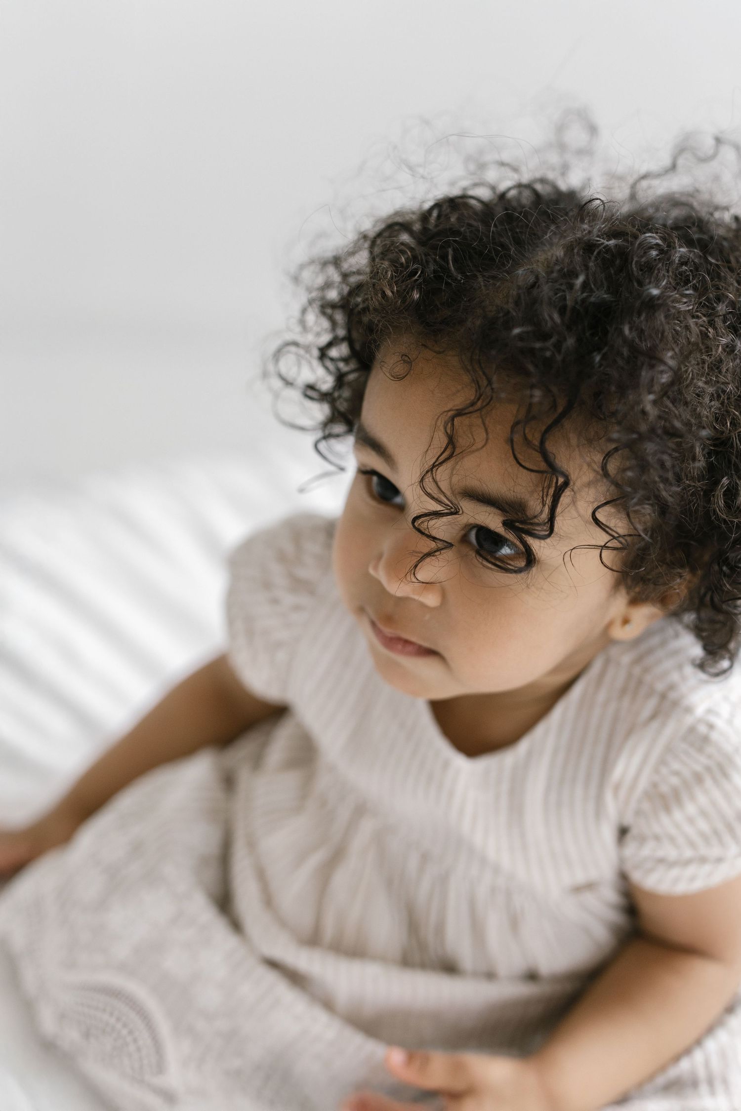 Curly-haired toddler in a striped linen outfit sitting on a white bed, looking away thoughtfully.