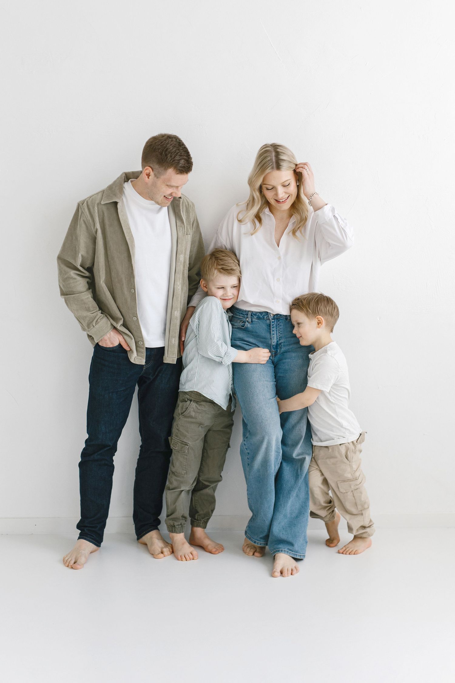 A family of four wearing casual clothing stands barefoot against a white backdrop, sharing a candid moment together.