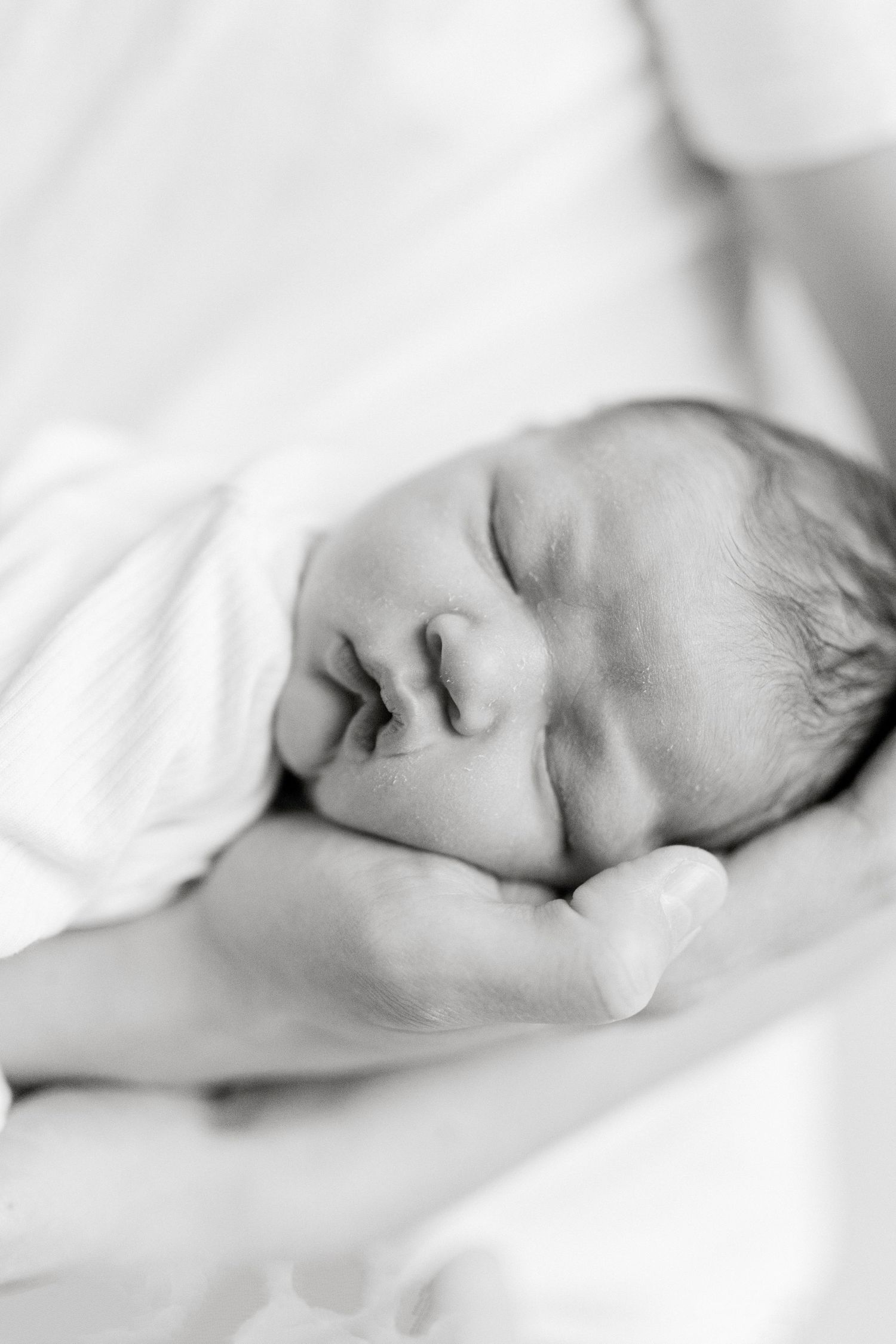 Black and white photo of a sleeping newborn baby cradled gently in an adult's hands, wrapped in a soft white blanket.