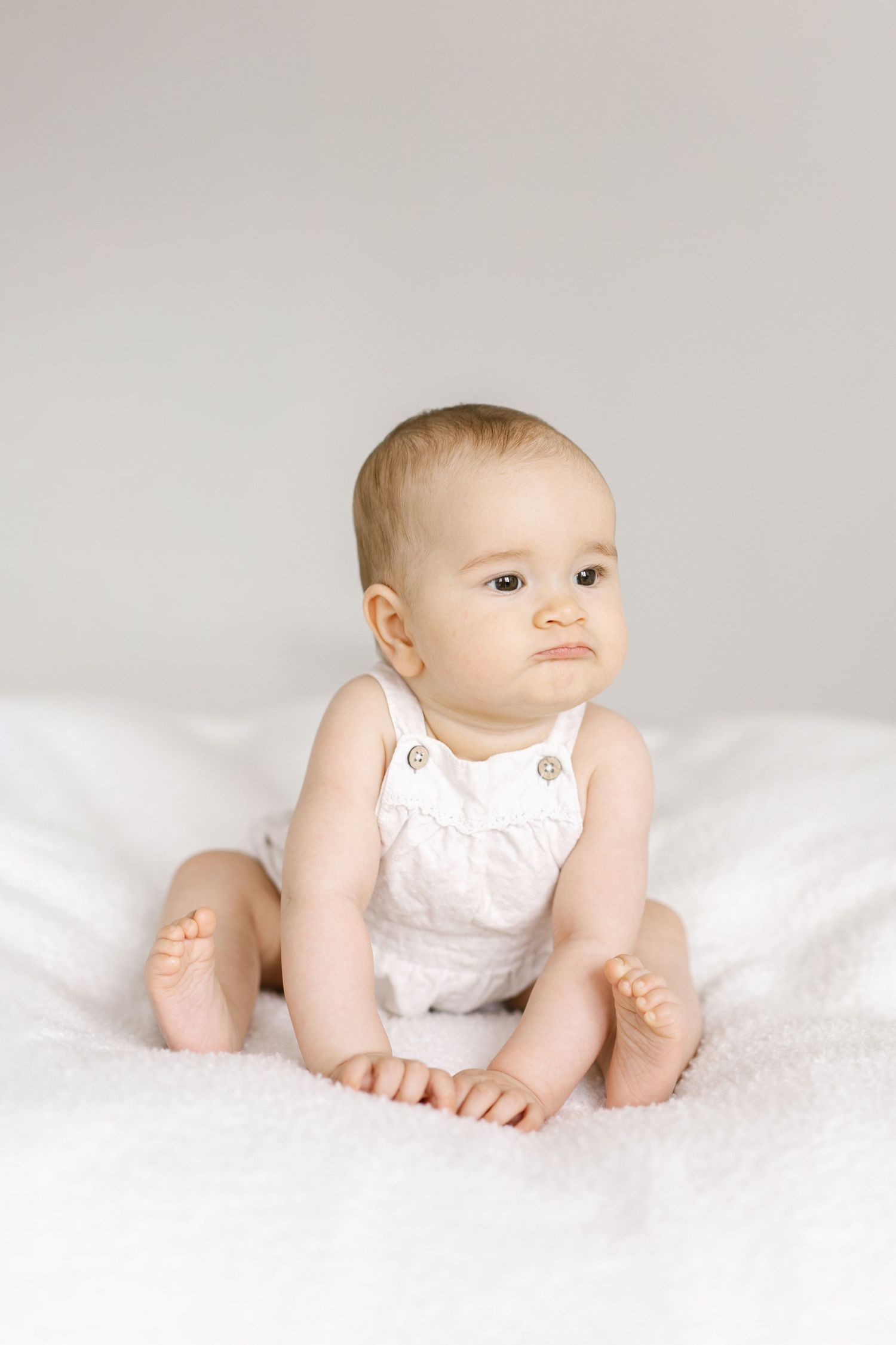 Adorable baby in white romper sitting on soft white blanket, looking sideways with curious expression.