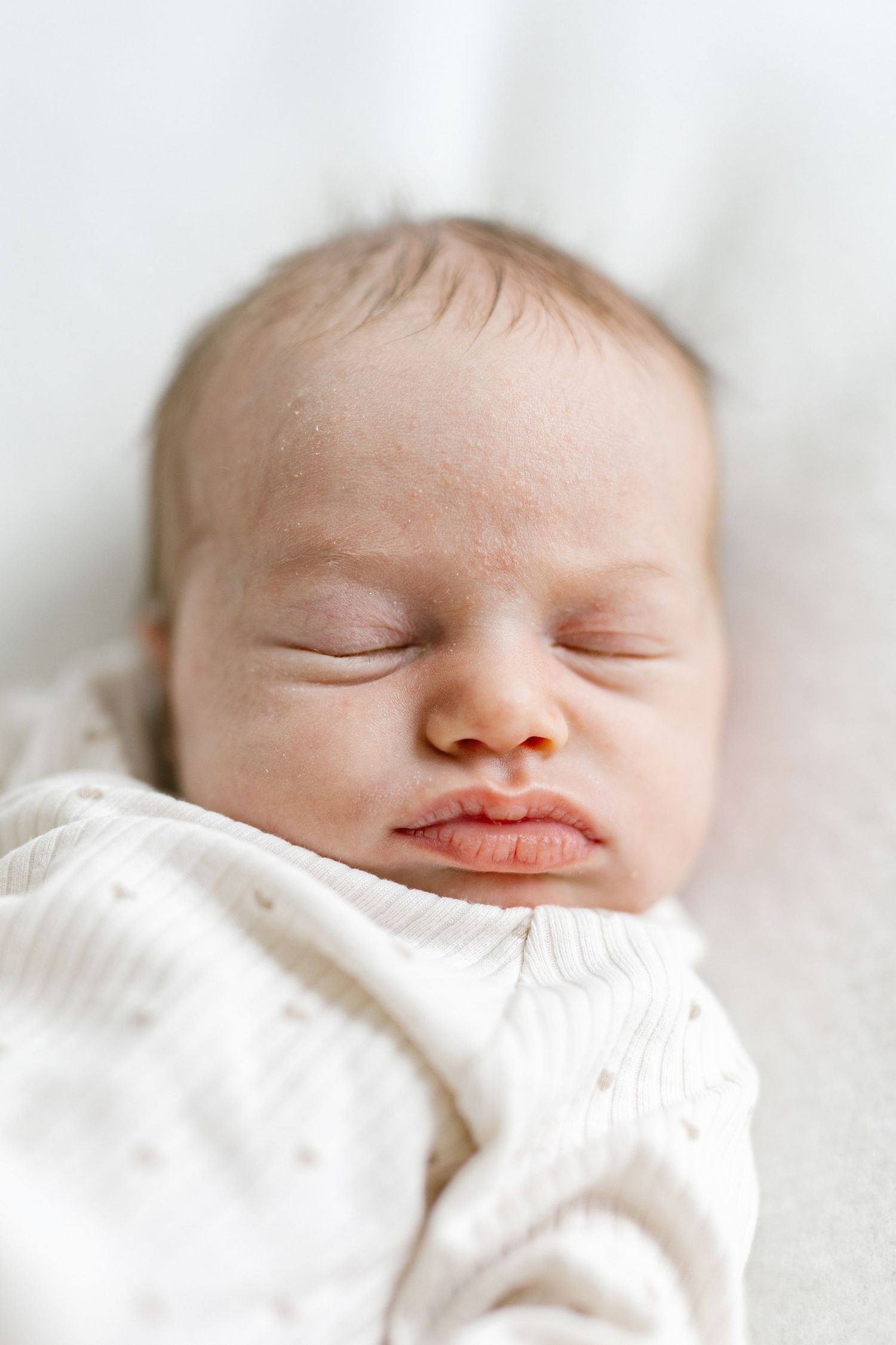 Peaceful sleeping newborn baby wrapped in a soft white knit blanket, with delicate features and tiny pursed lips.