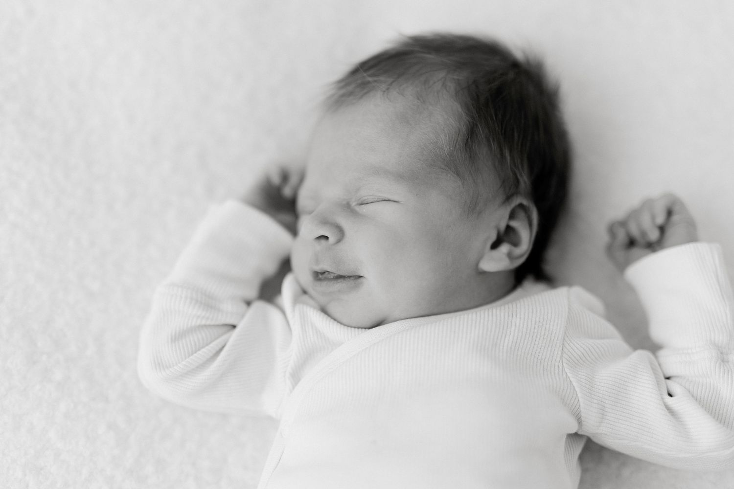 Black and white newborn baby sleeping peacefully on soft white blanket, arms raised, wearing white onesie.