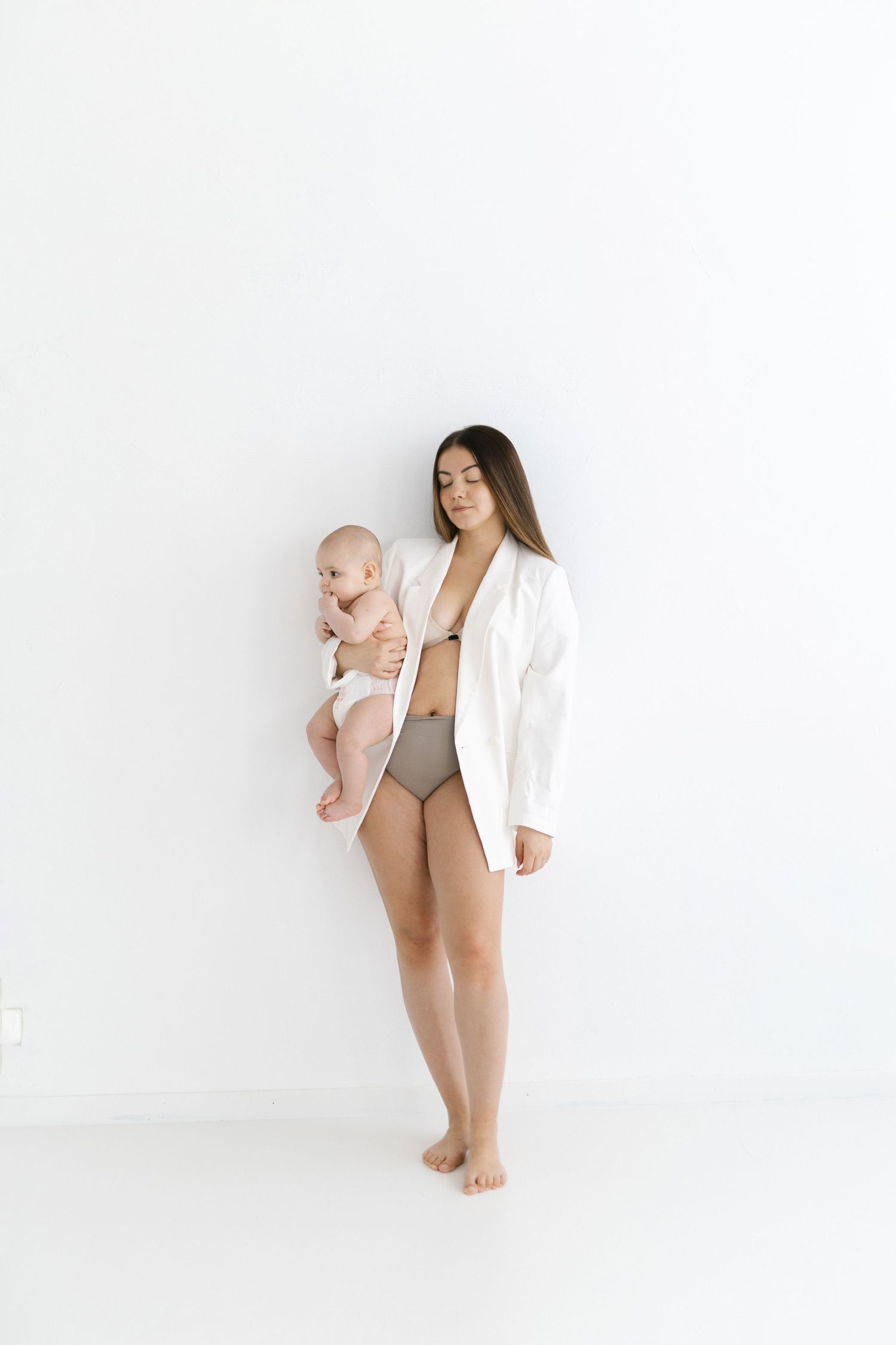 A young mother in white blazer and underwear holds her newborn baby against a white background.