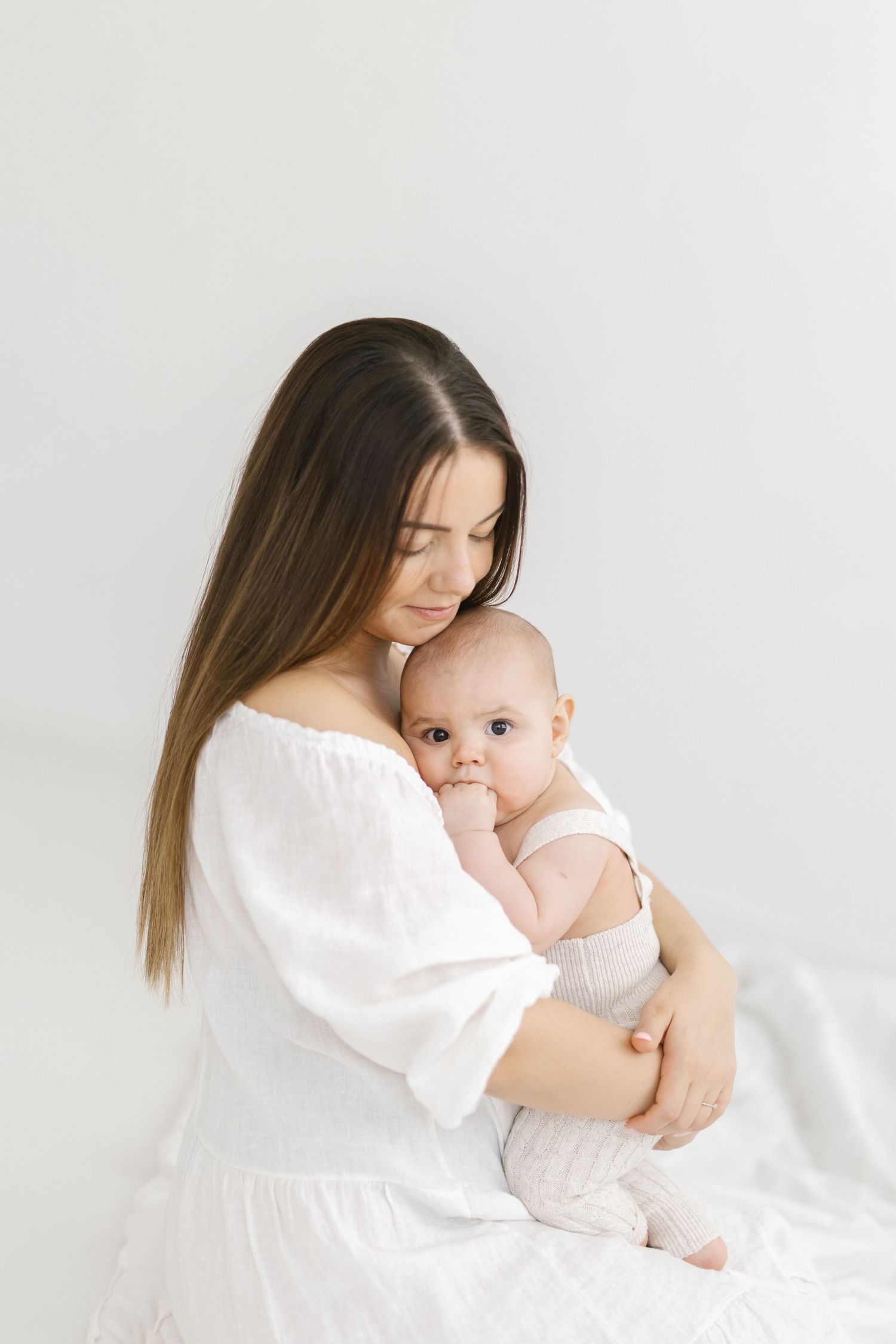 A young mother in a white off-shoulder dress tenderly holds her alert baby in a bright, minimalist white setting.