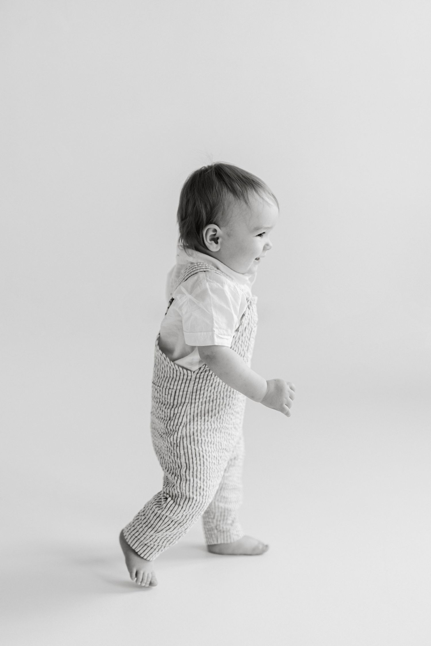 Baby in striped overalls practicing walking in minimalist white setting.