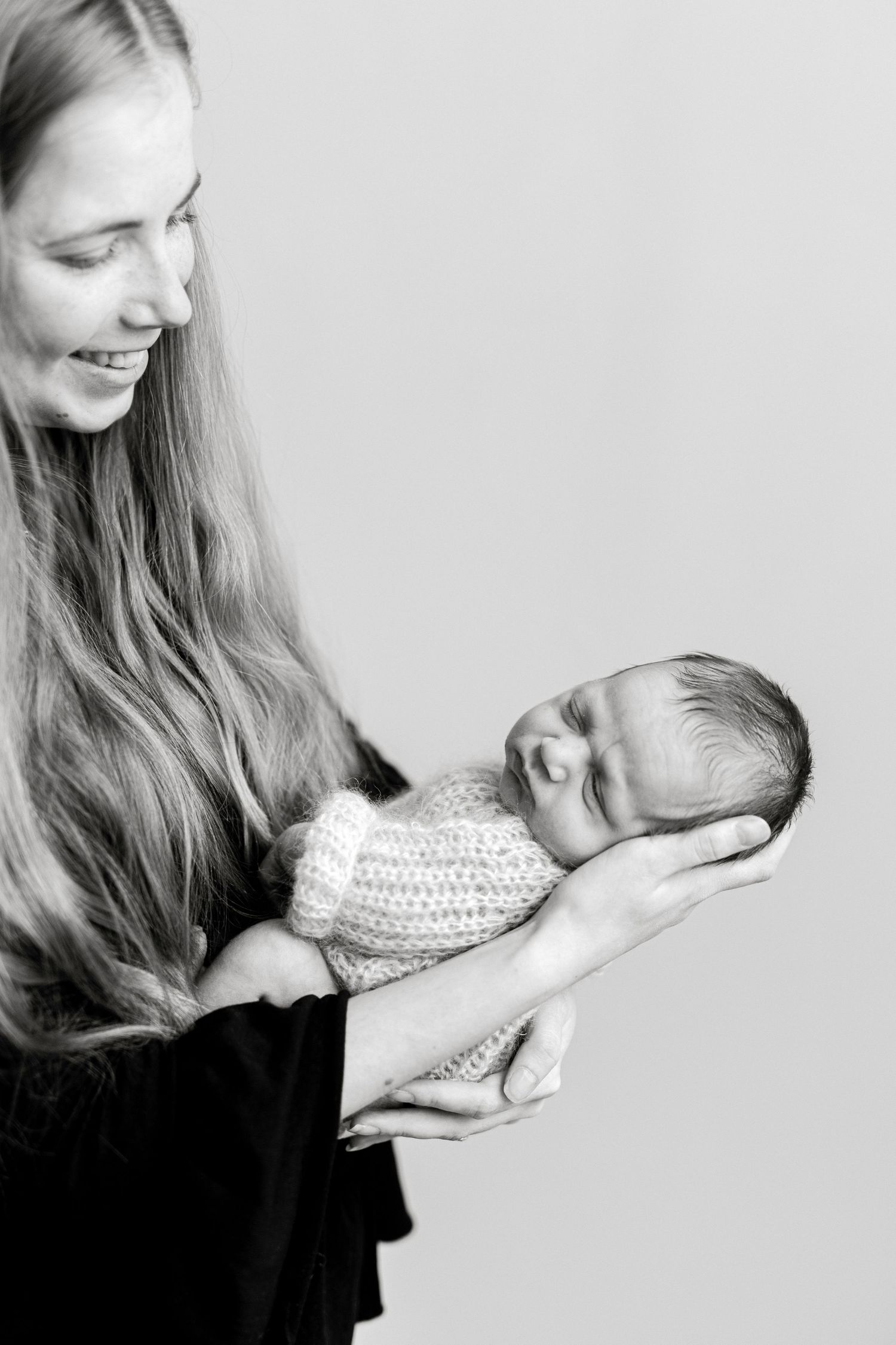 Smiling mother with long hair holds sleeping newborn baby wrapped in knit blanket, black and white portrait.
