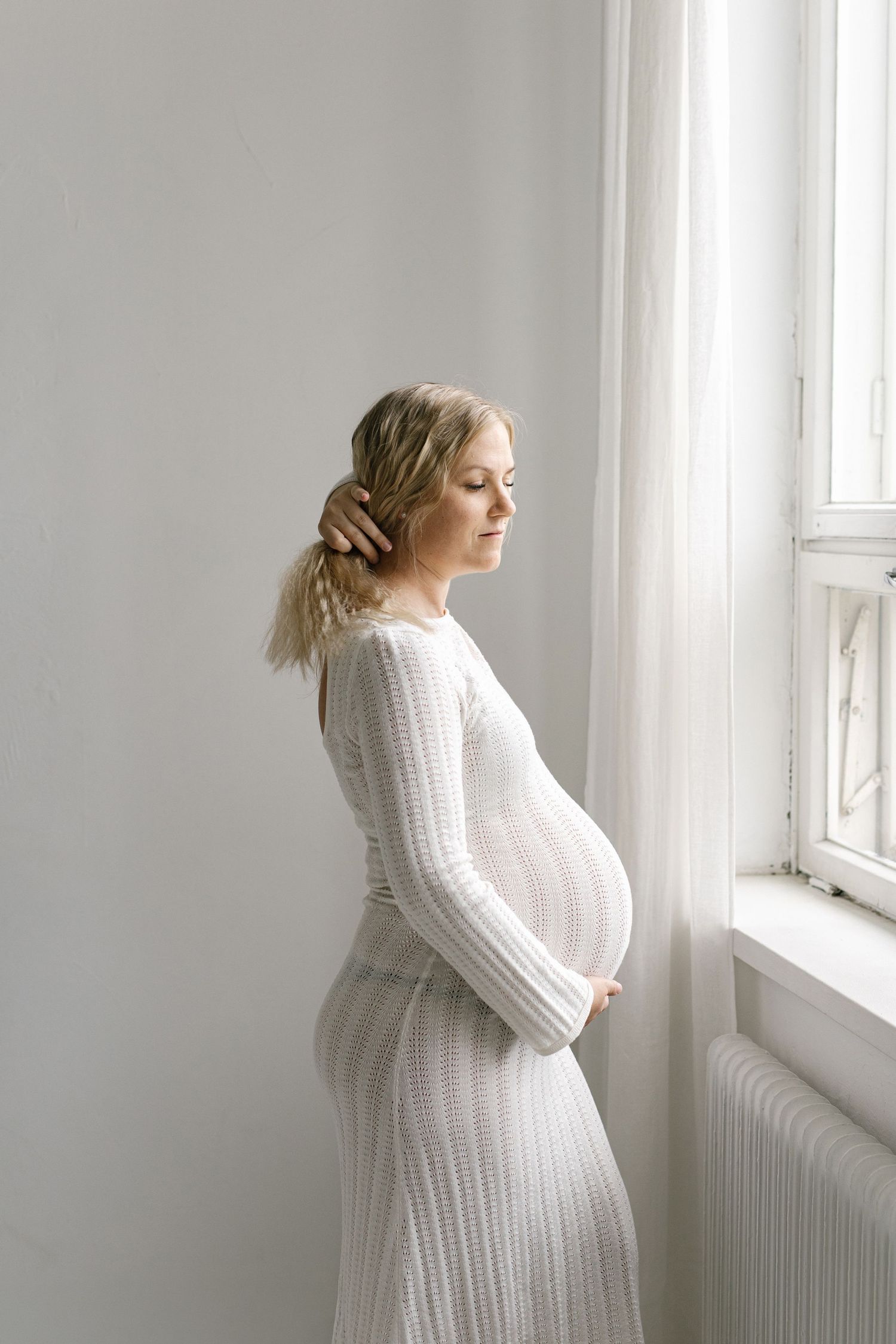 Pregnant woman in white knit dress cradling baby bump while gazing out a bright window.