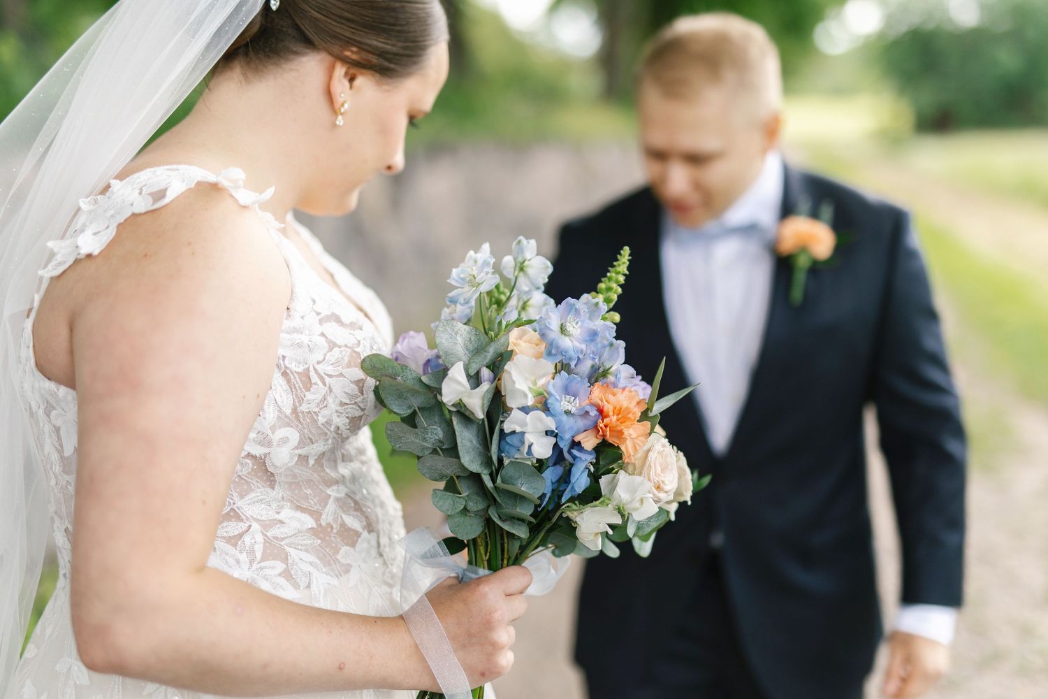 Bride in lace wedding dress holding blue and peach bouquet with groom in navy suit during outdoor wedding ceremony.