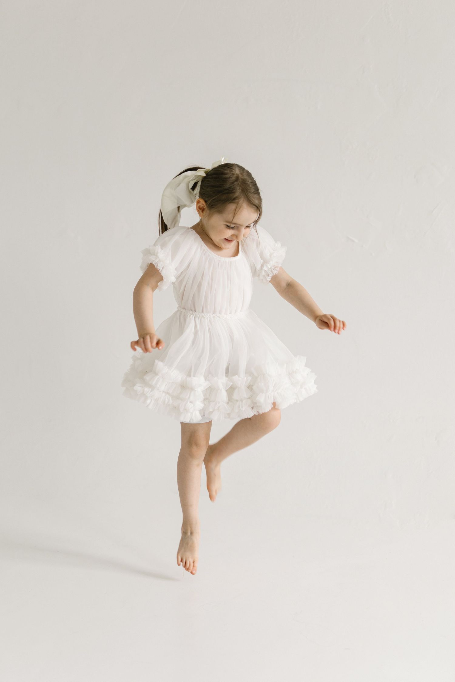 A young girl in a white ruffled dress and bow dances barefoot on one leg against a white background.
