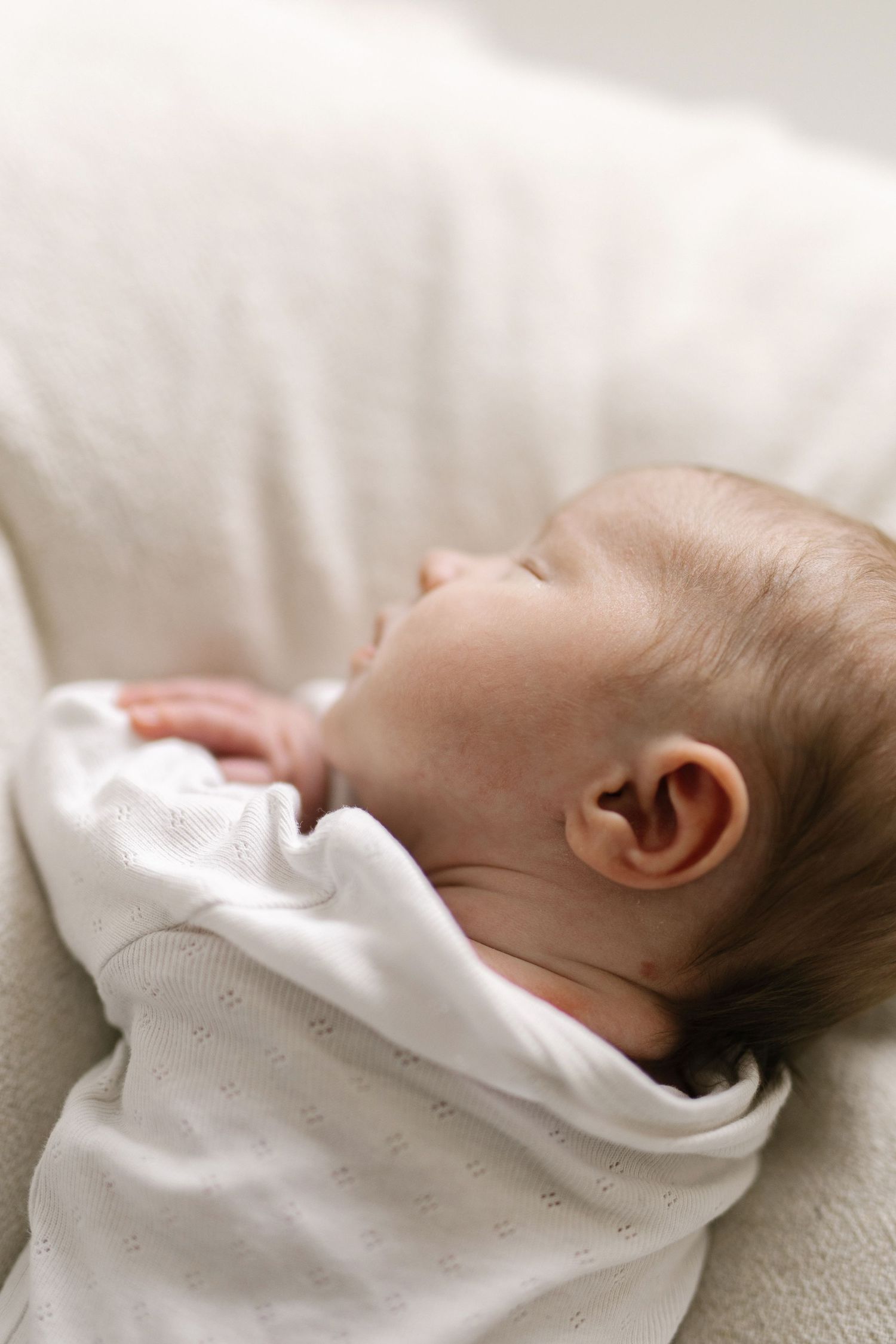 A peaceful newborn baby sleeping soundly, swaddled in a soft white blanket against a light background.