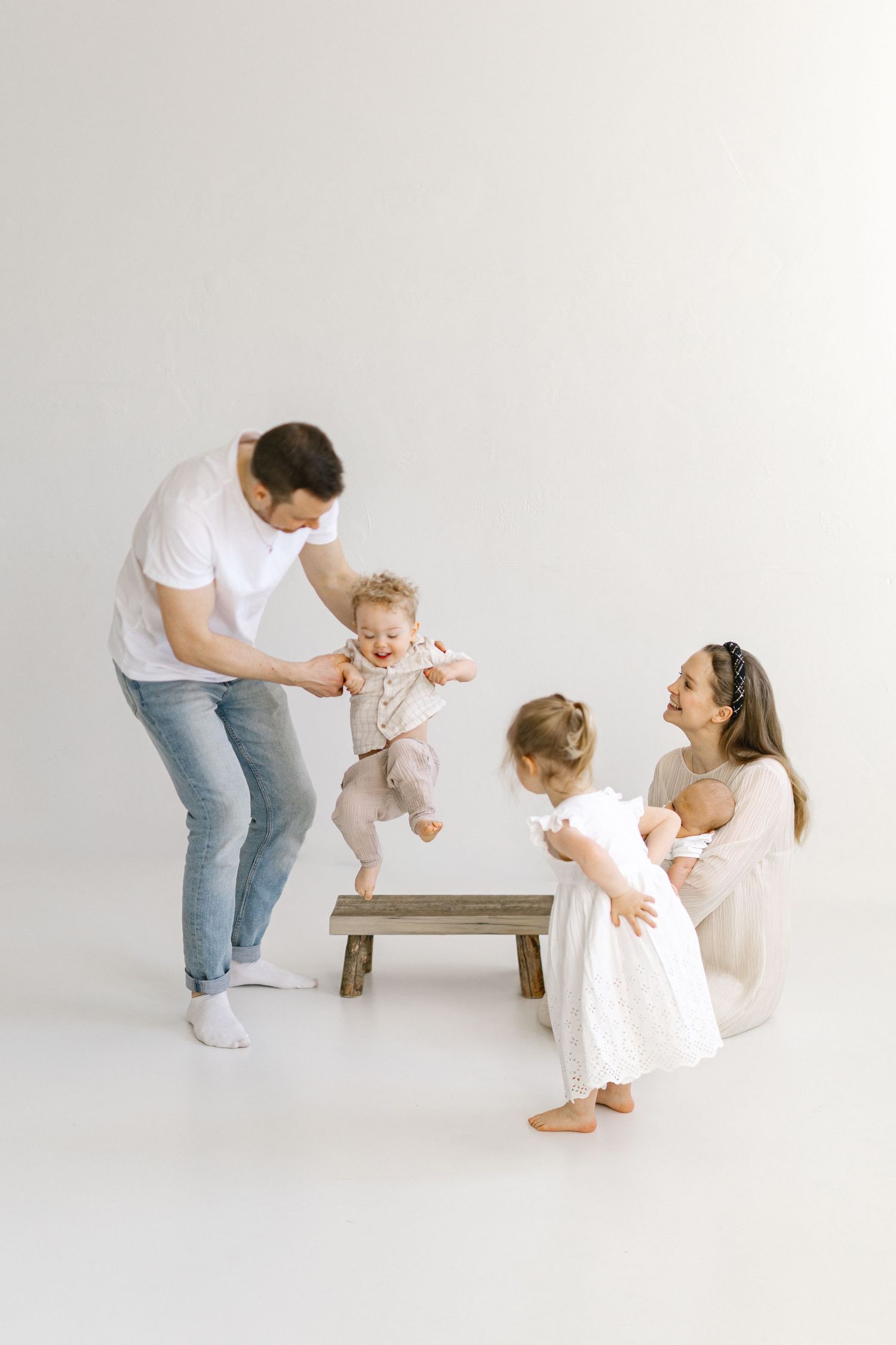 A happy family with a dad, mom, toddler girl, and baby enjoying a playful moment together in a bright white studio.