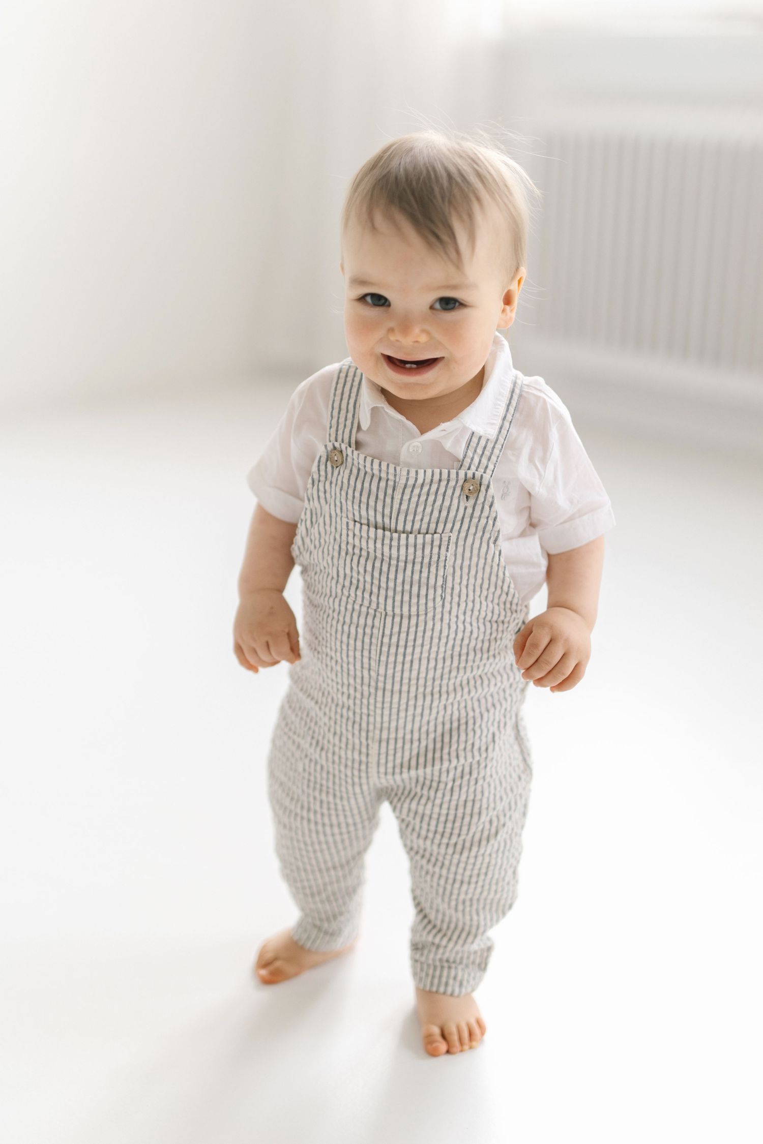 Baby in striped overalls standing and smiling in white room.