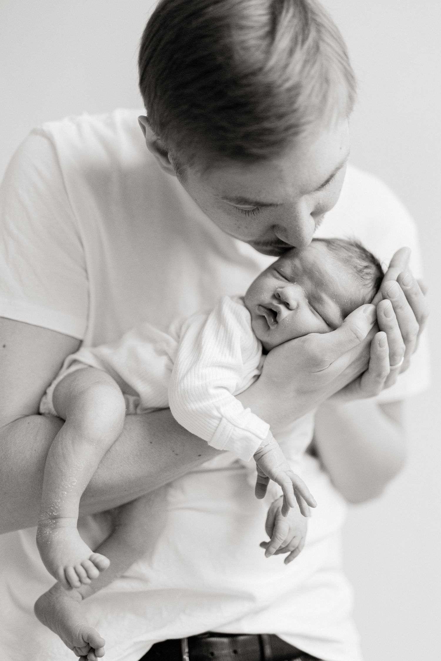 Black and white photo of a father tenderly kissing his newborn baby while cradling the infant in both hands.