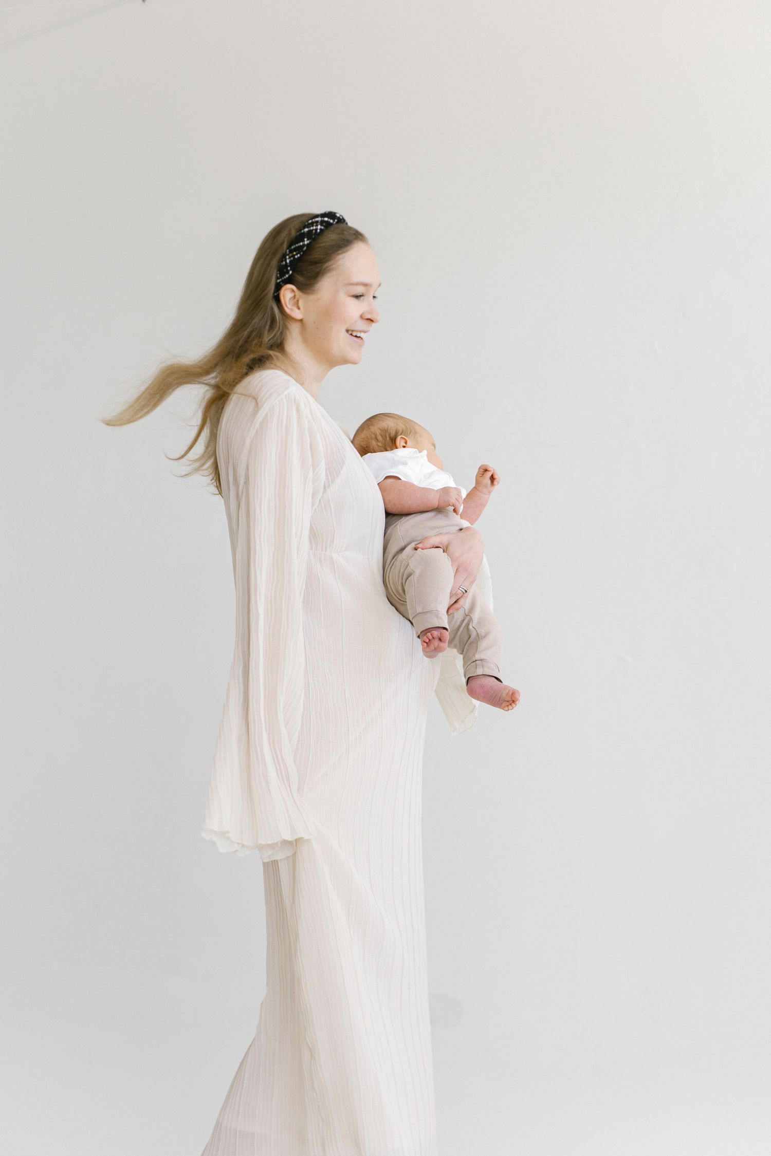 Smiling mother in white dress holding newborn baby against a white background.