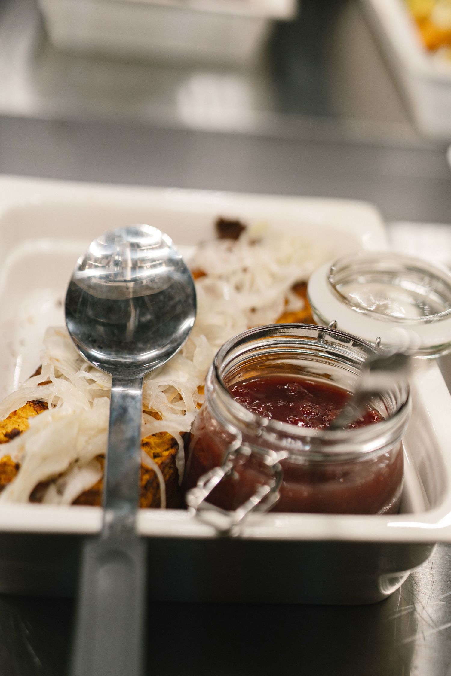 Glass jar of red sauce with a spoon beside noodles and grilled food on a white tray.