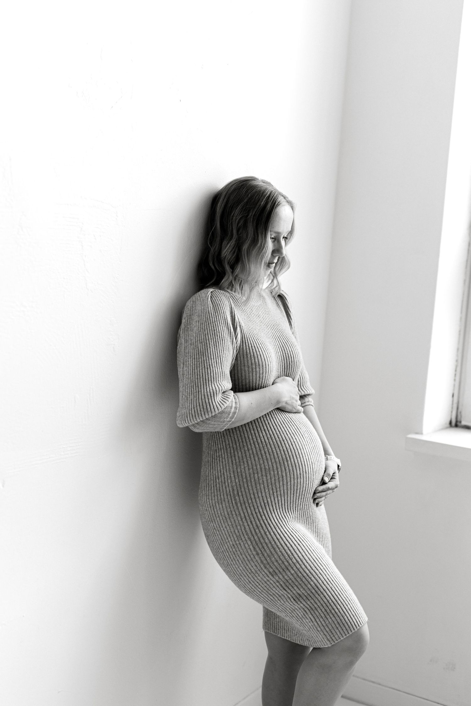 Black and white photo of a pregnant woman in a ribbed dress cradling her baby bump while leaning against a white wall.