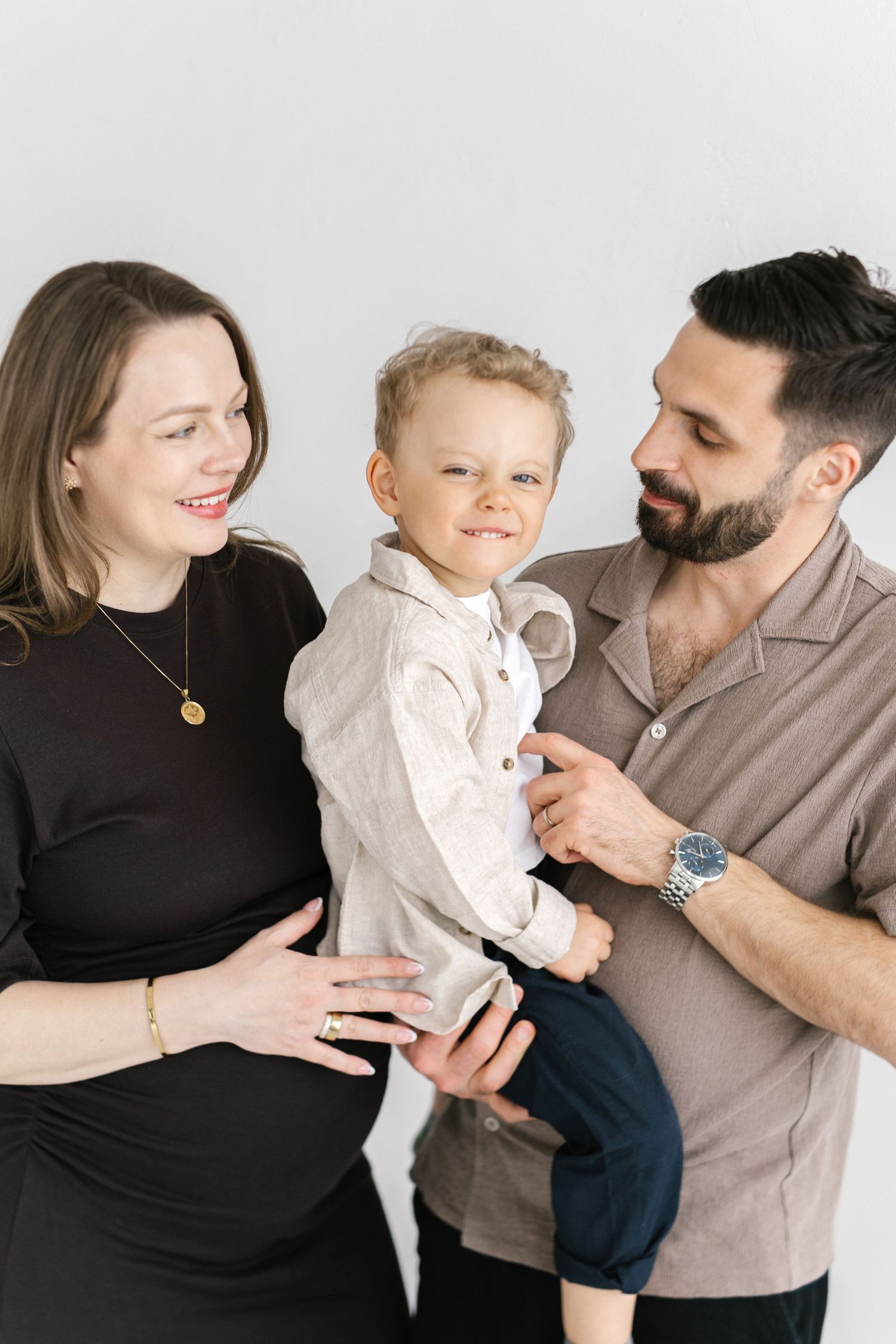A happy family portrait with a pregnant woman, bearded man, and smiling toddler boy against a white background.