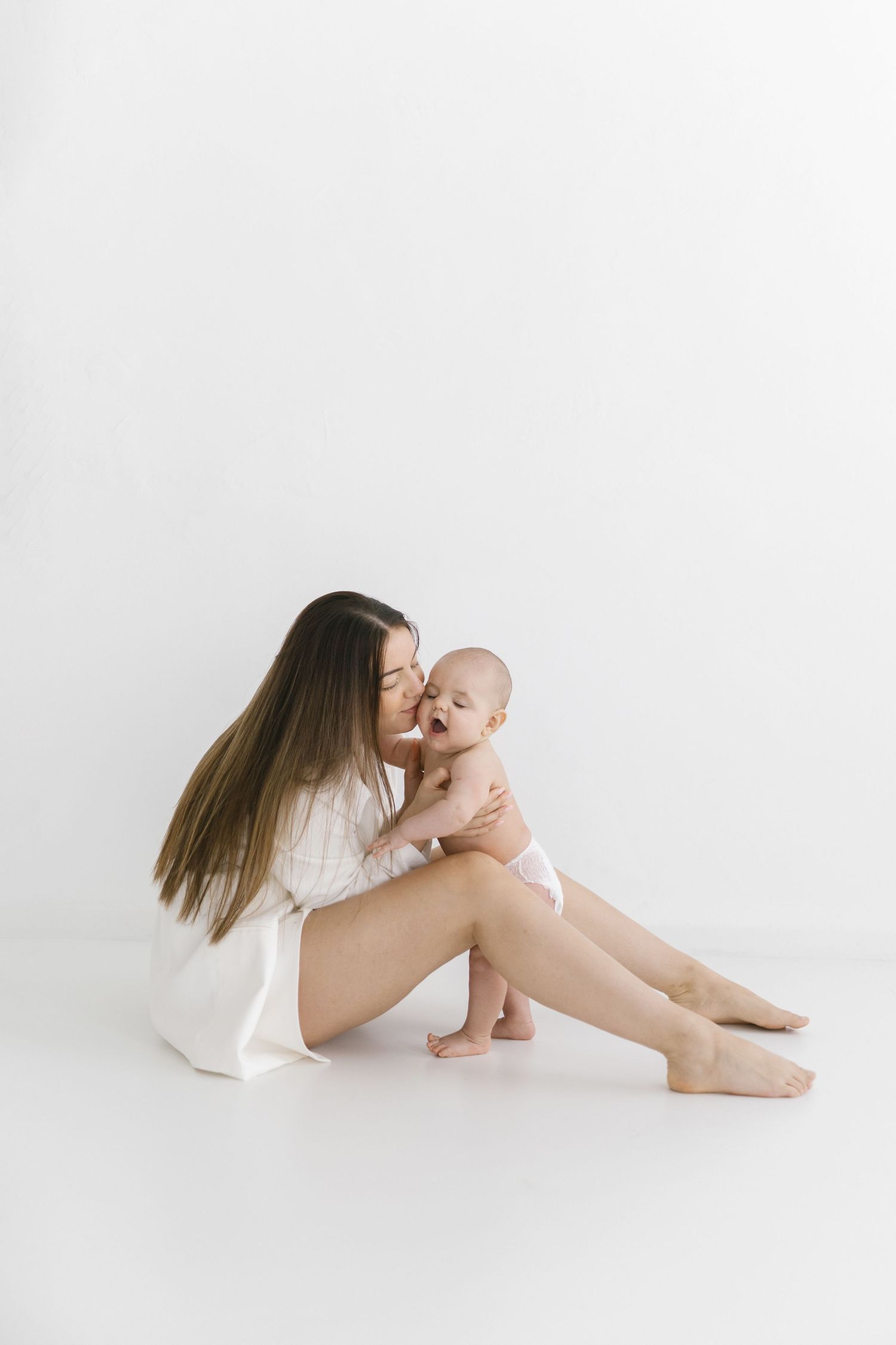 A young mother in a white dress sits on a white floor, lovingly kissing her laughing baby in a diaper.