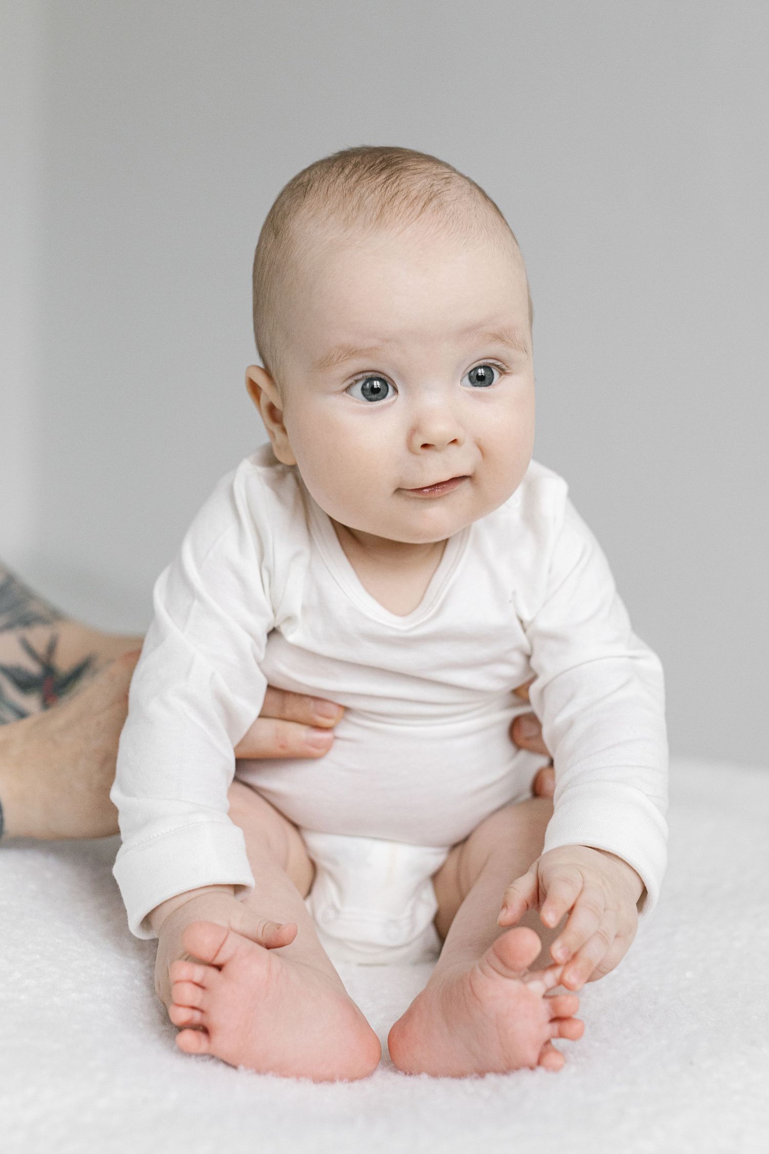 Adorable baby in white onesie sitting upright on soft white surface, looking at camera with bright curious eyes.
