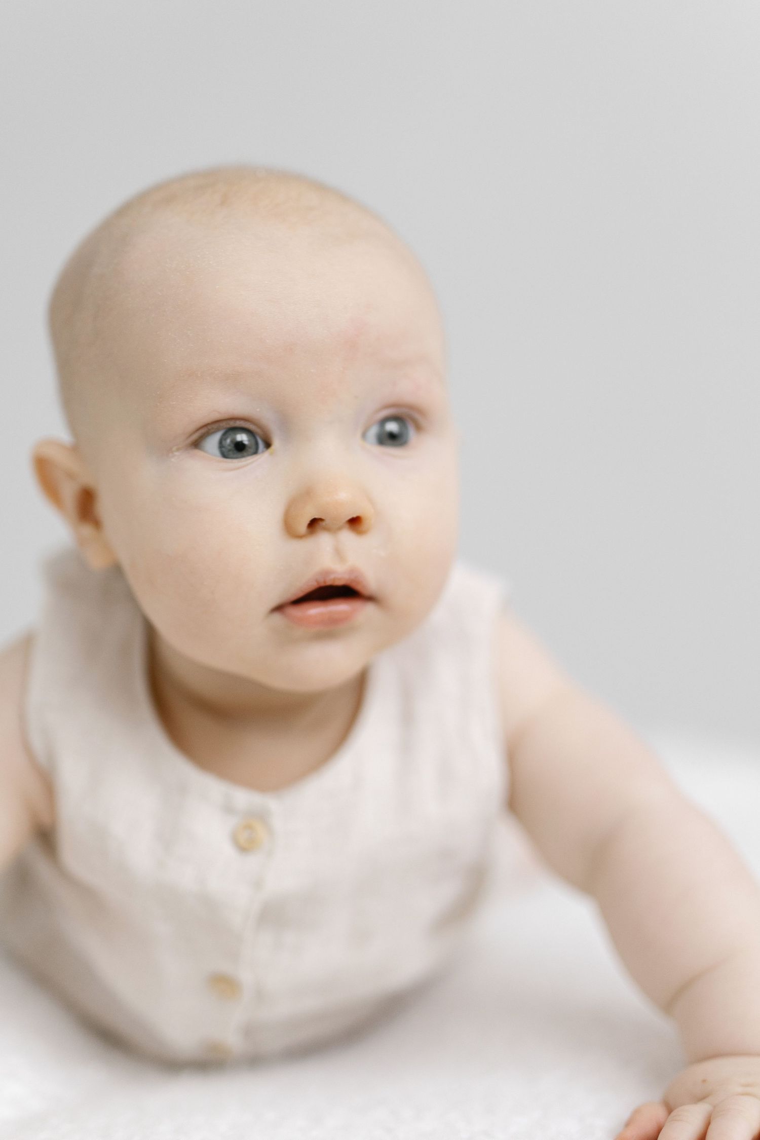 Cute baby with blue eyes wearing a cream linen romper lying on a white surface.