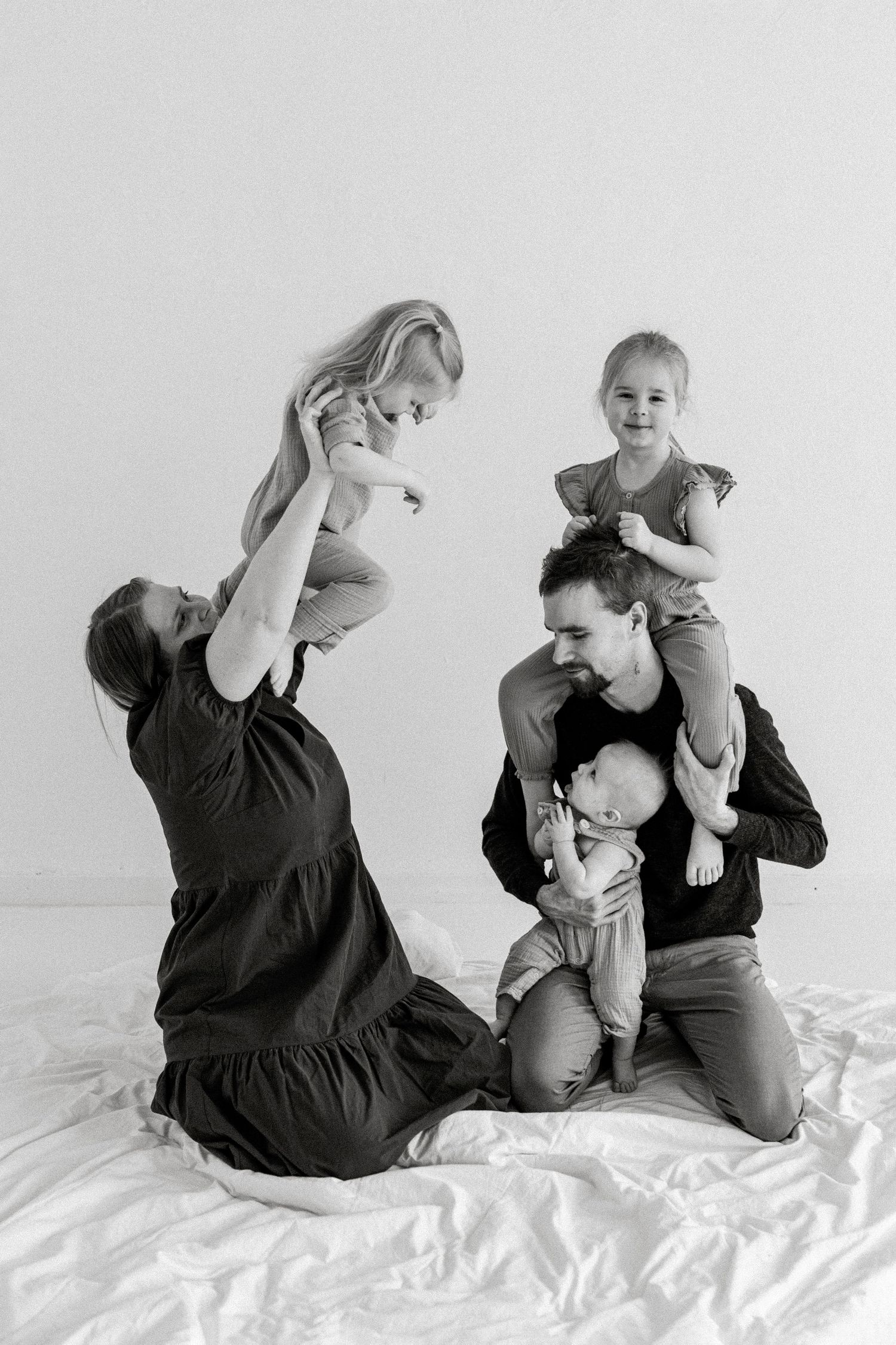 Black and white family photo of parents playfully lifting three young children on a white bed.