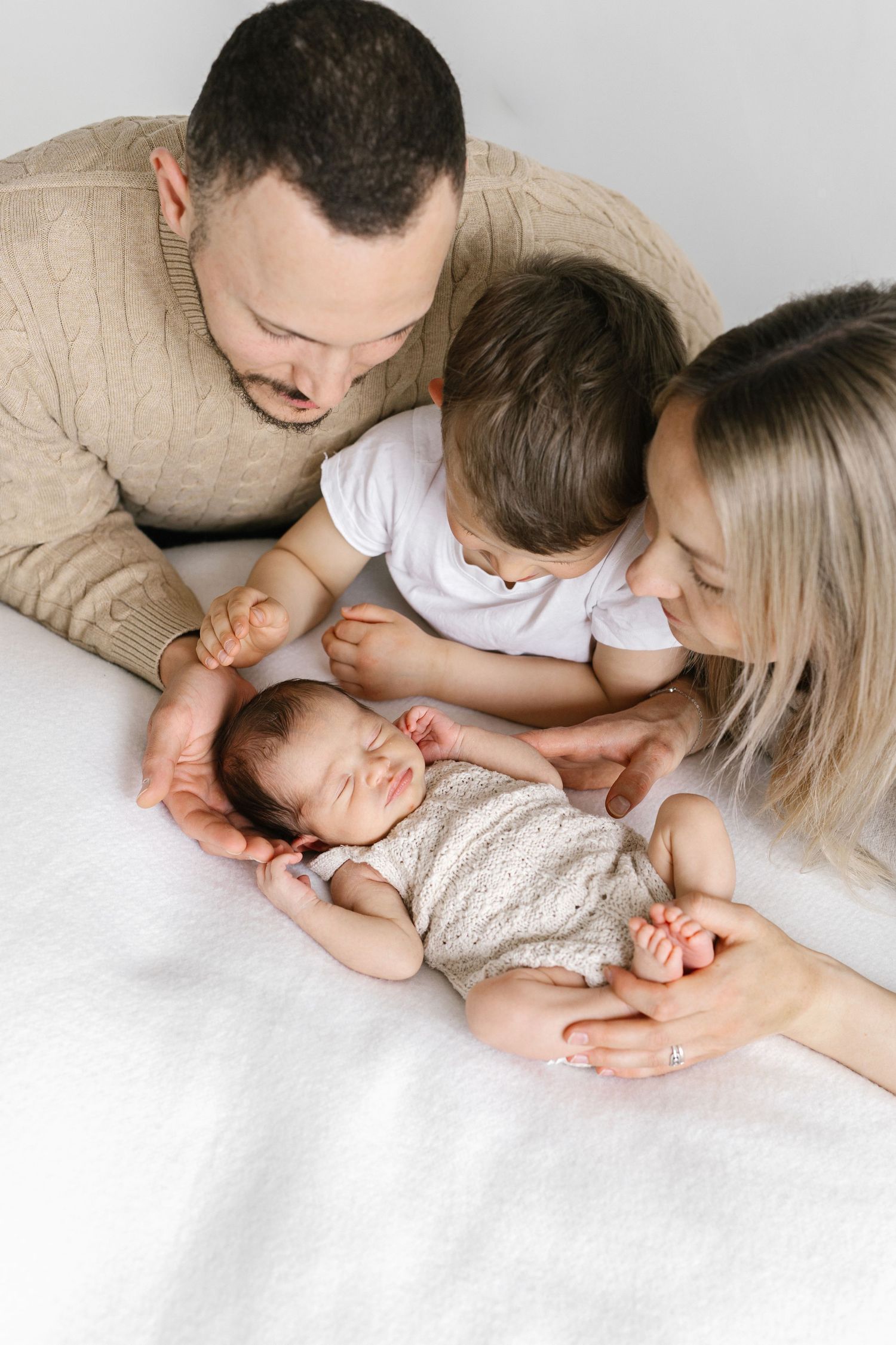 A family of four admires their sleeping newborn baby, with parents and toddler gathered lovingly on a white bed.