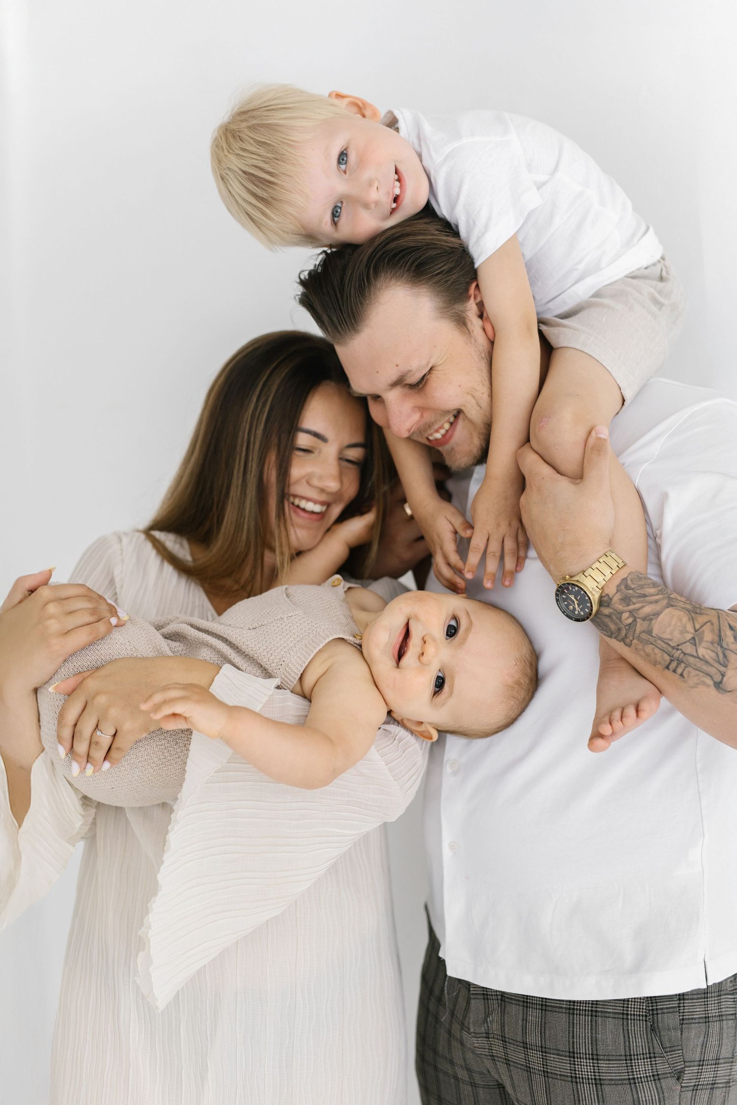 Happy family with two children, parents laughing as toddler sits on dad's shoulders and baby rests in arms.