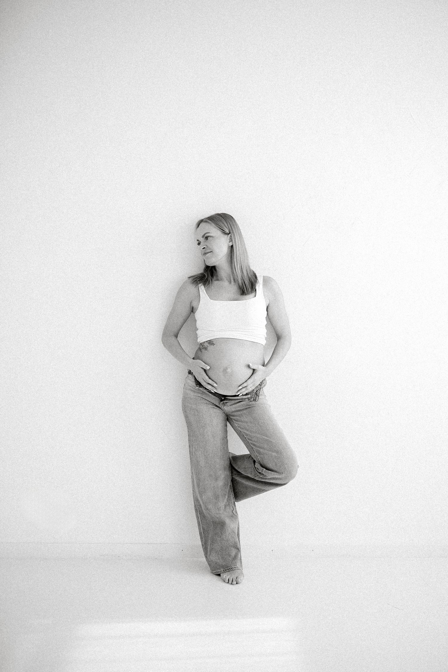 Artistic maternity photos with woman in white crop top and baggy jeans against white wall.