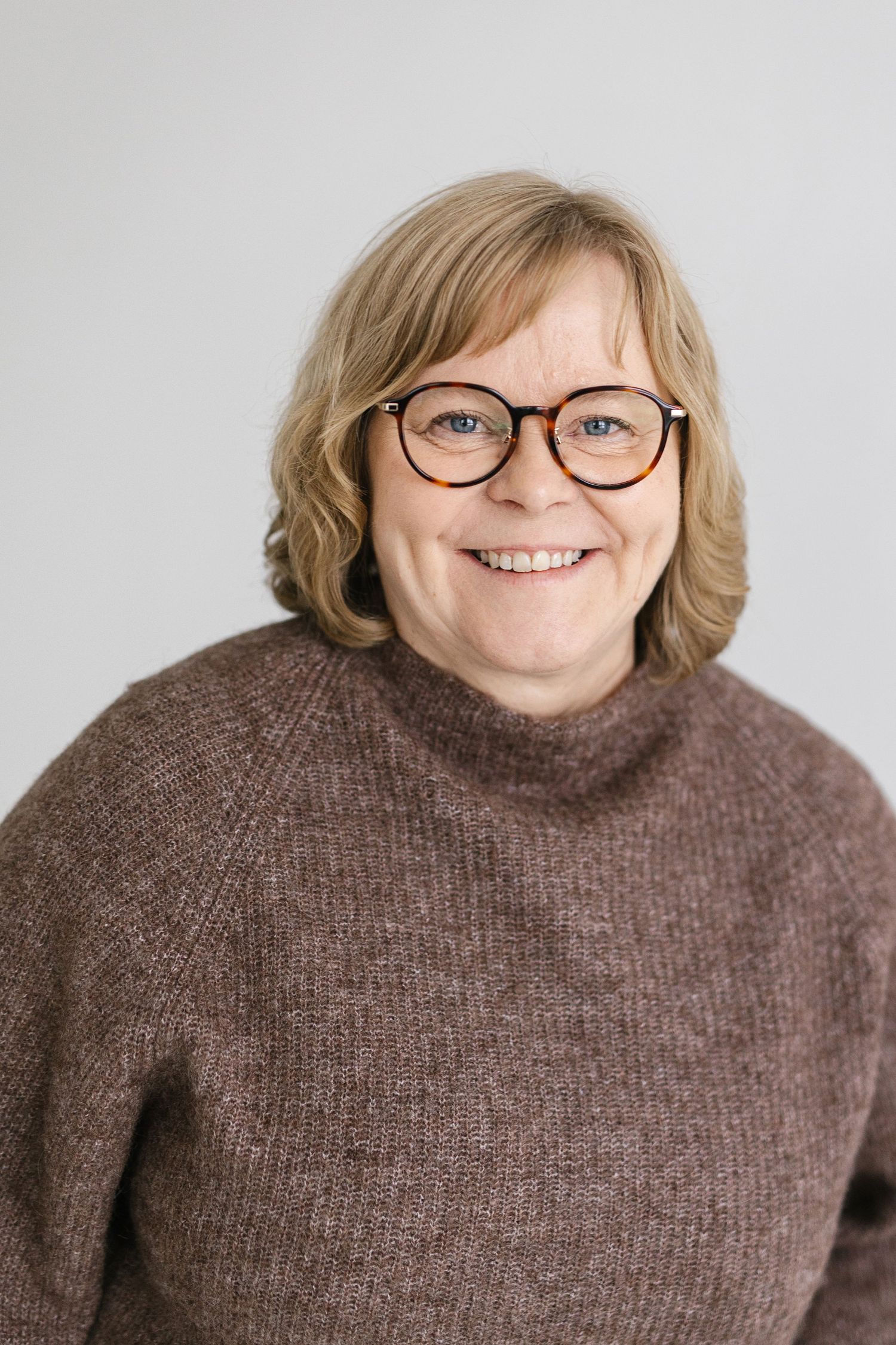 A person wearing glasses and a brown sweater smiles warmly against a light background.