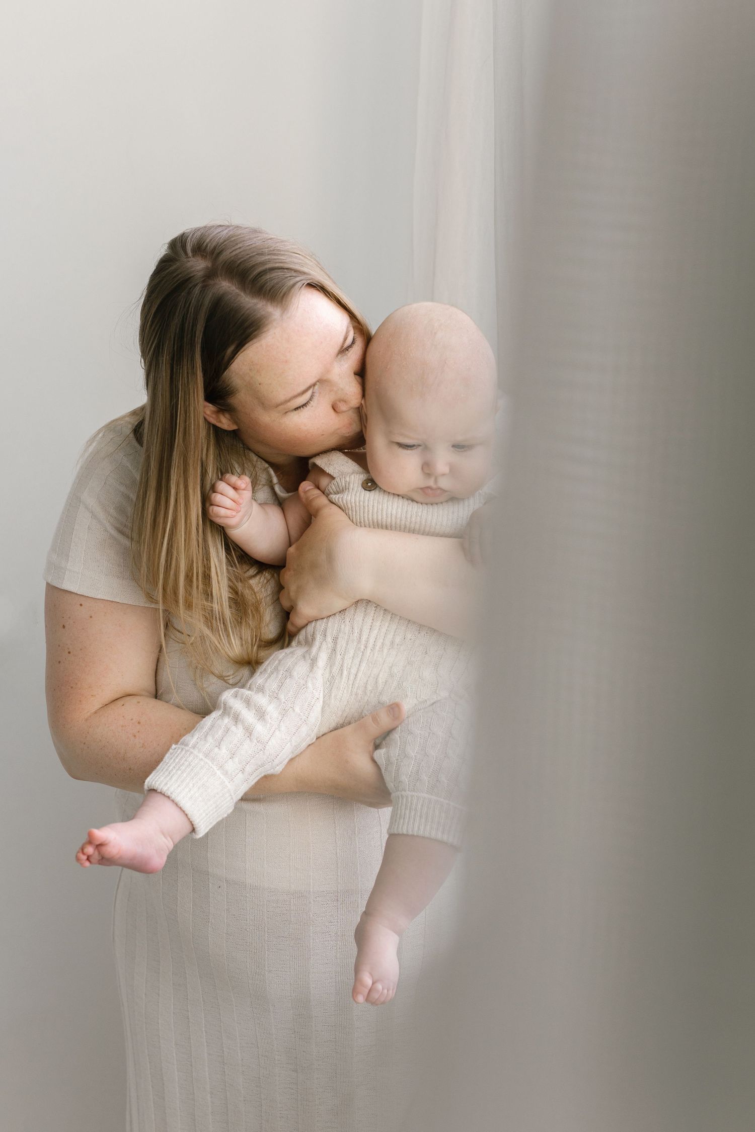 A mother in a cream dress tenderly kisses her newborn baby wrapped in a white knit outfit.