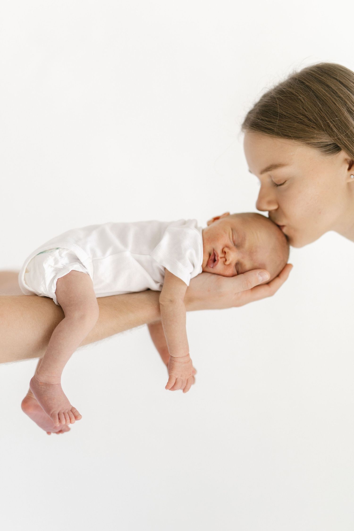 A tender mother gently kisses her sleeping newborn baby dressed in white, cradled in loving hands against a white background.
