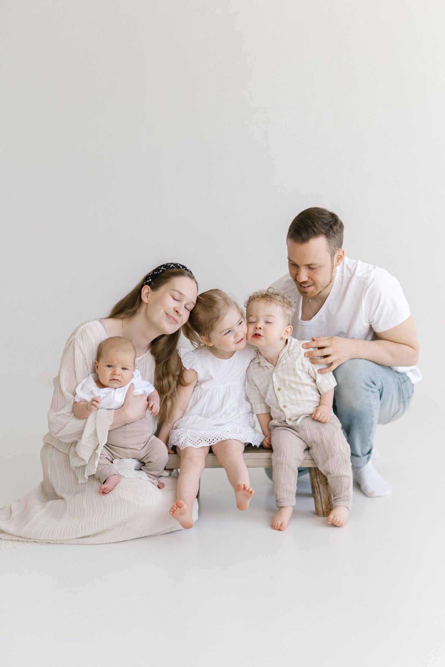 A happy family of five poses in a bright studio, with parents and three young children dressed in soft neutral tones.