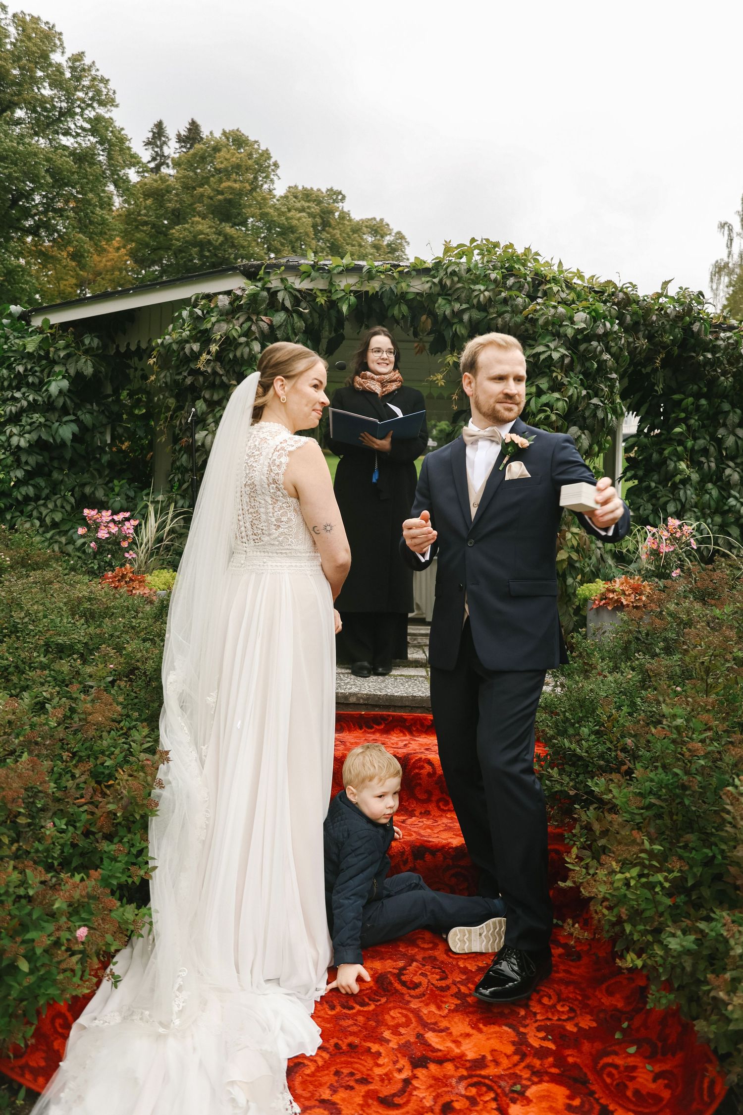 Bride and groom exchange vows at outdoor garden wedding while young child plays at their feet.