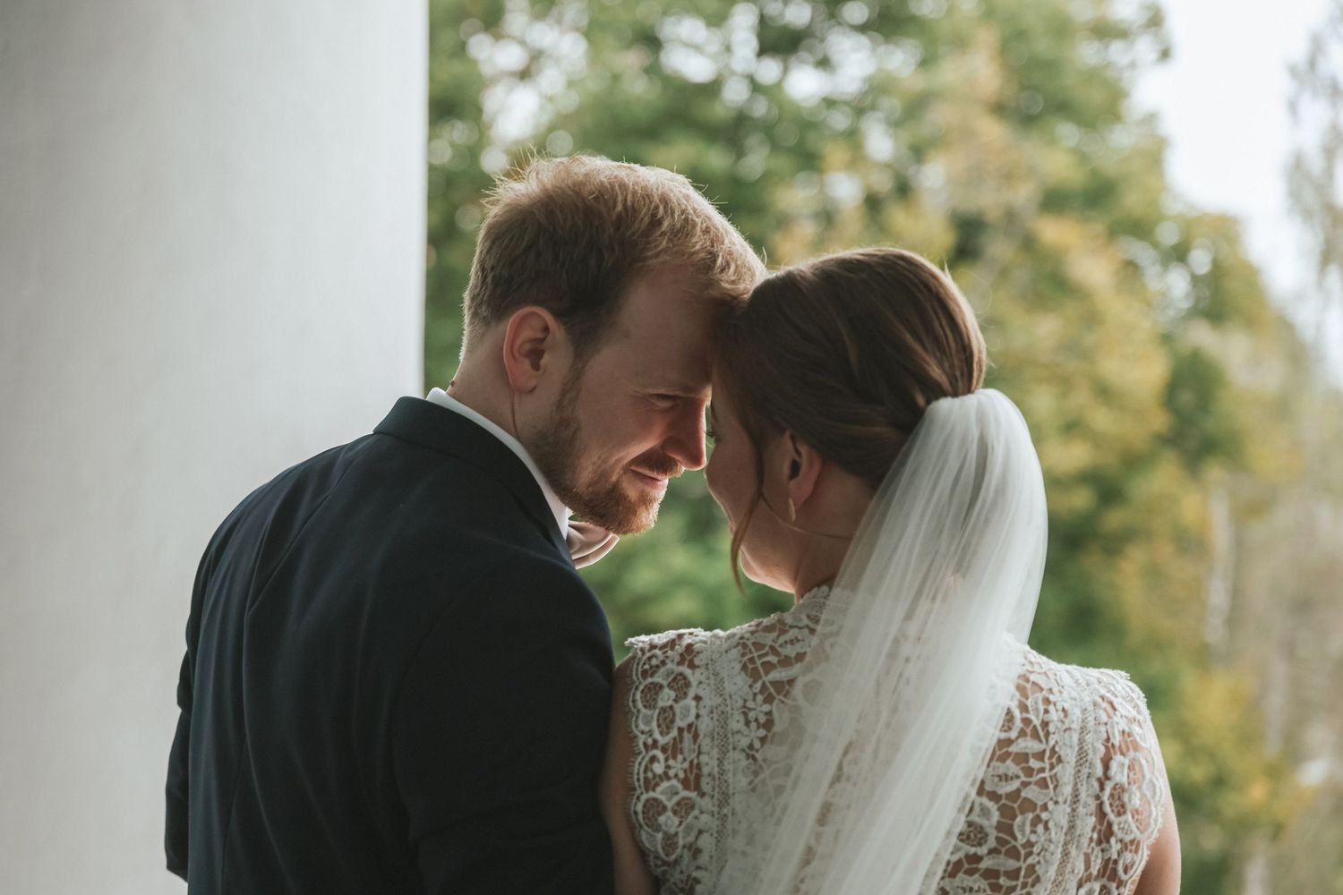Romantic bride and groom touching foreheads tenderly in a lace wedding dress and black suit outdoors.