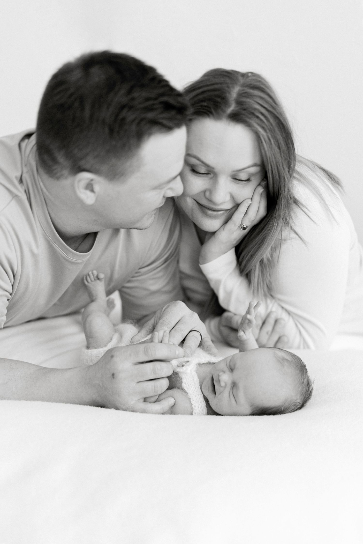 Black and white photo of smiling parents lying down with their newborn baby.