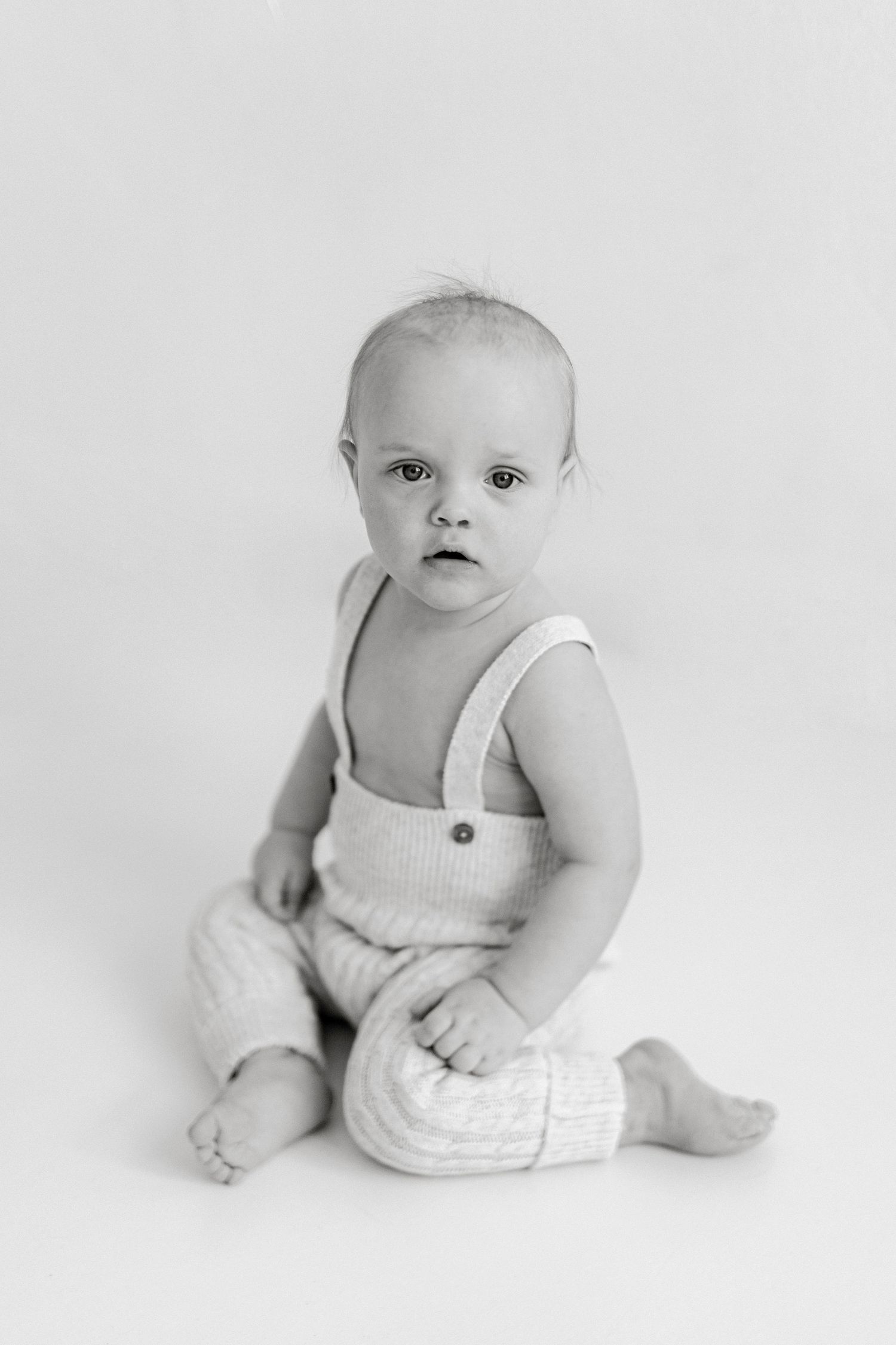 Black and white portrait of a cute baby sitting upright wearing suspender overalls on a white background.
