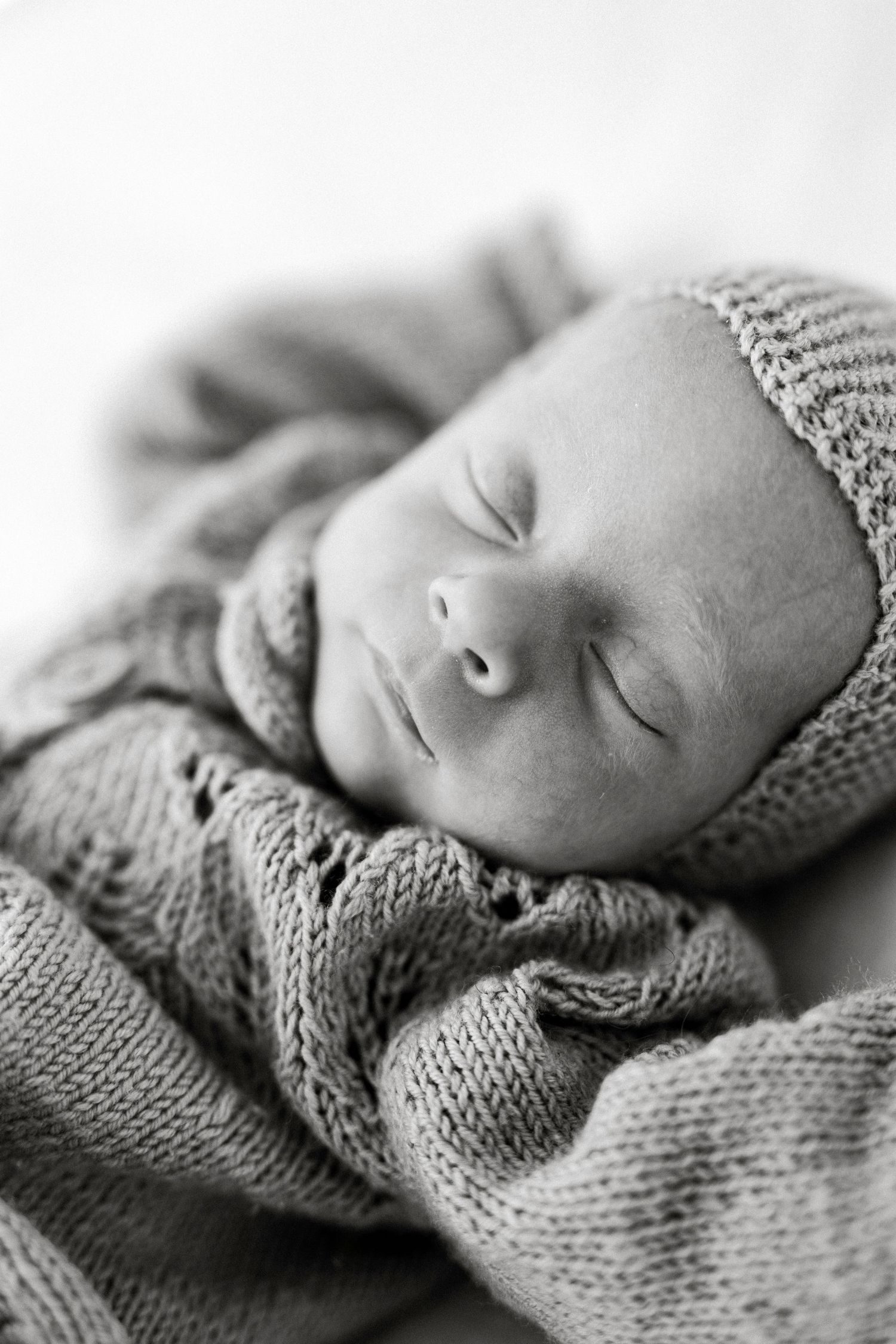 Black and white photo of a sleeping newborn baby wrapped in a soft knit blanket and matching hat.