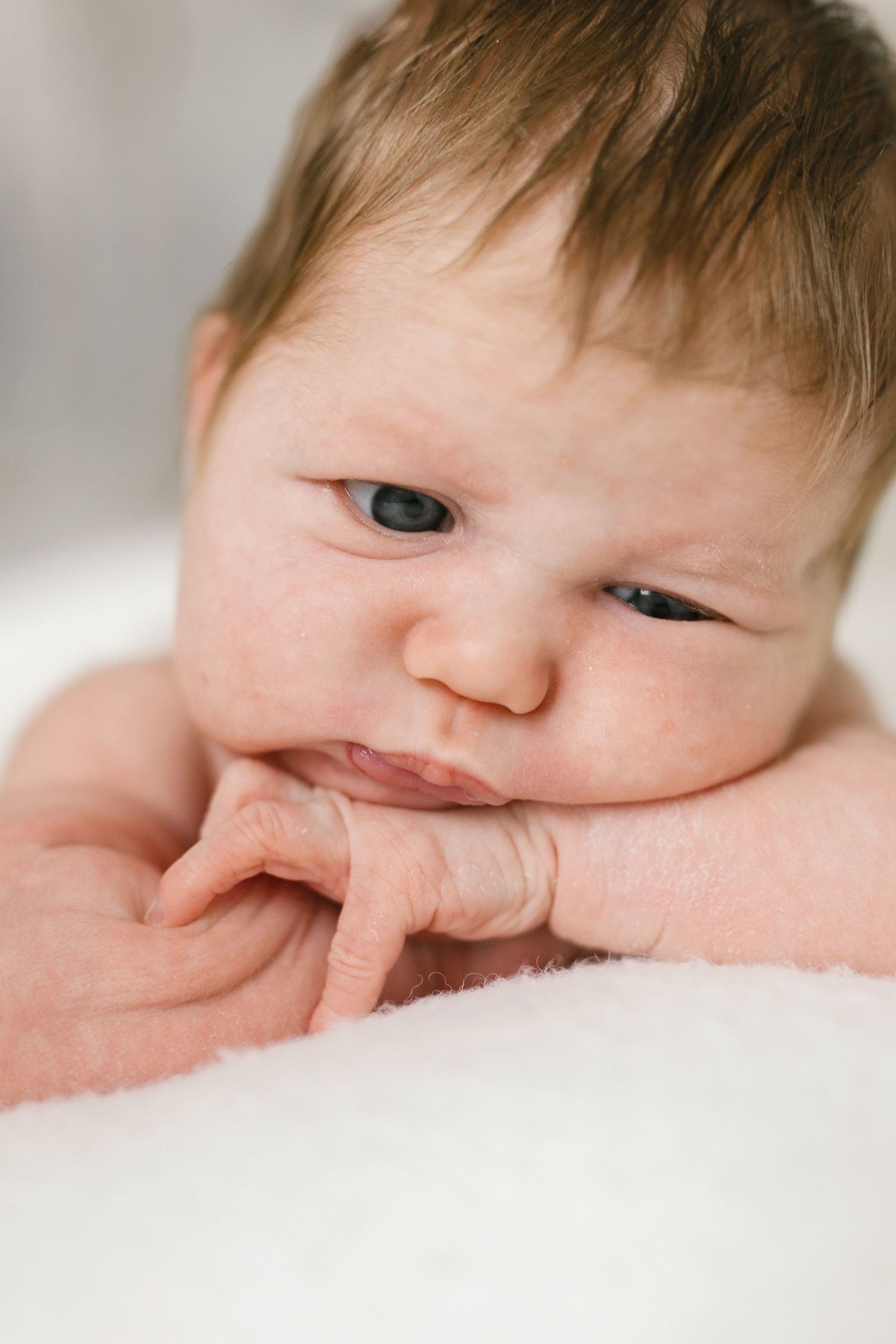 Newborn baby with light brown hair rests chin on tiny folded hands with a grumpy, skeptical expression on a soft white blanket.