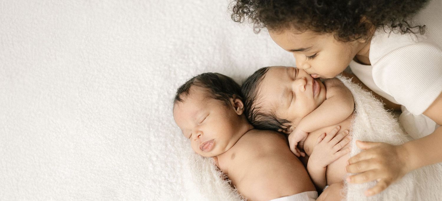Older sibling gently kissing newborn twins wrapped in white blanket in a sweet family portrait.