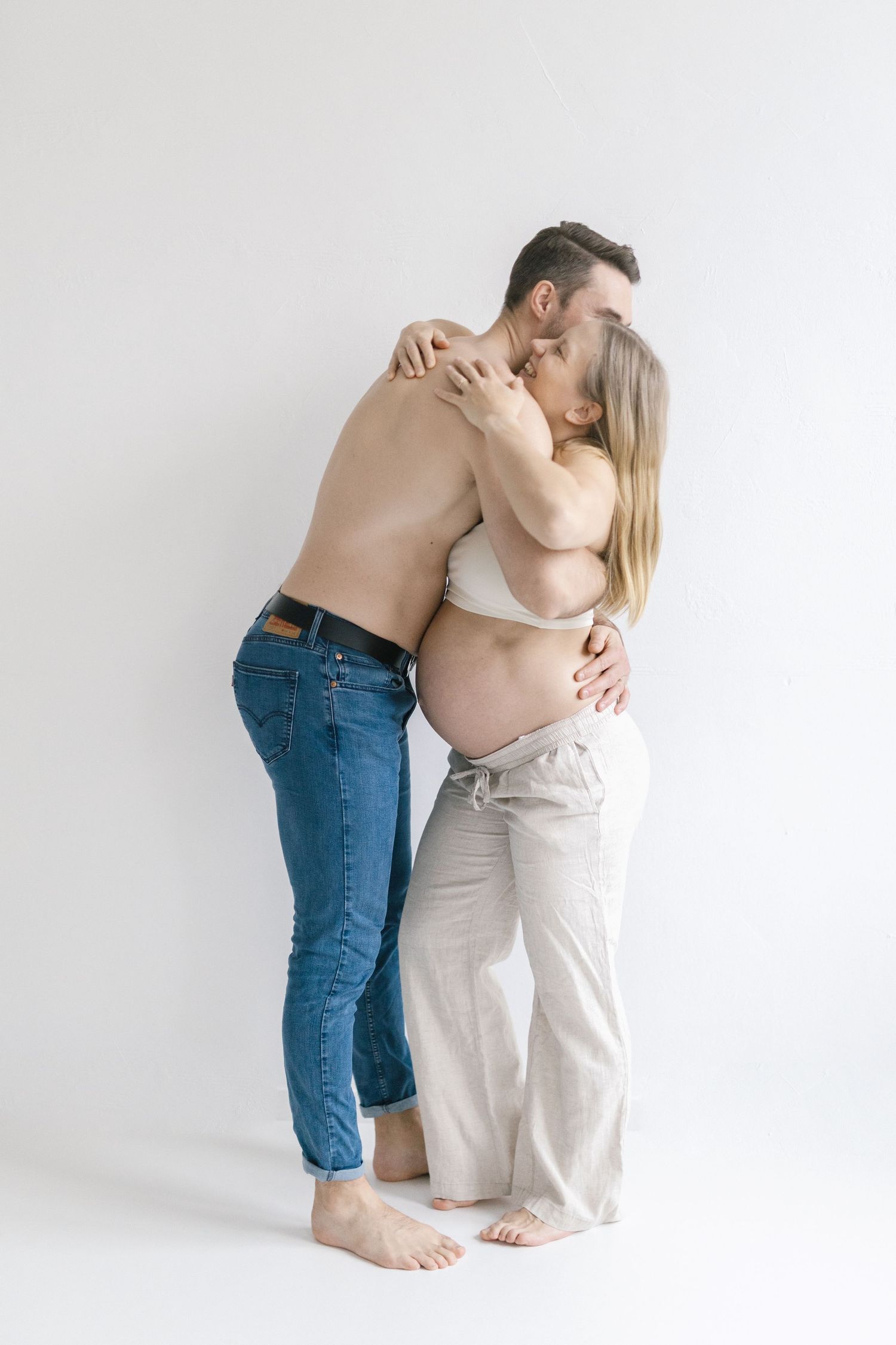 Shirtless man embracing pregnant woman in white bra and gray pants, sharing a tender kiss on white background.