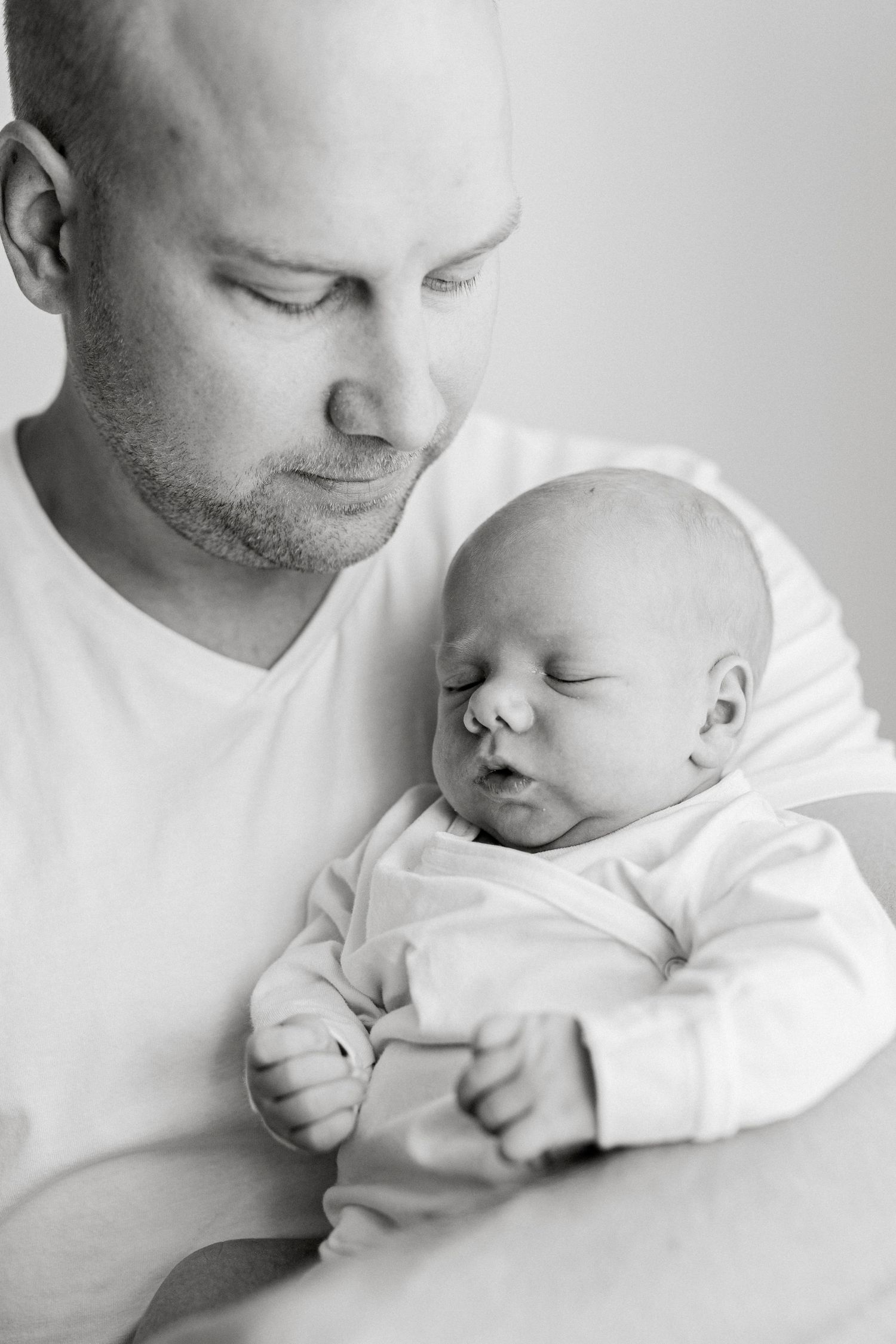 Black and white photo of a bald father tenderly holding his sleeping newborn baby dressed in white.