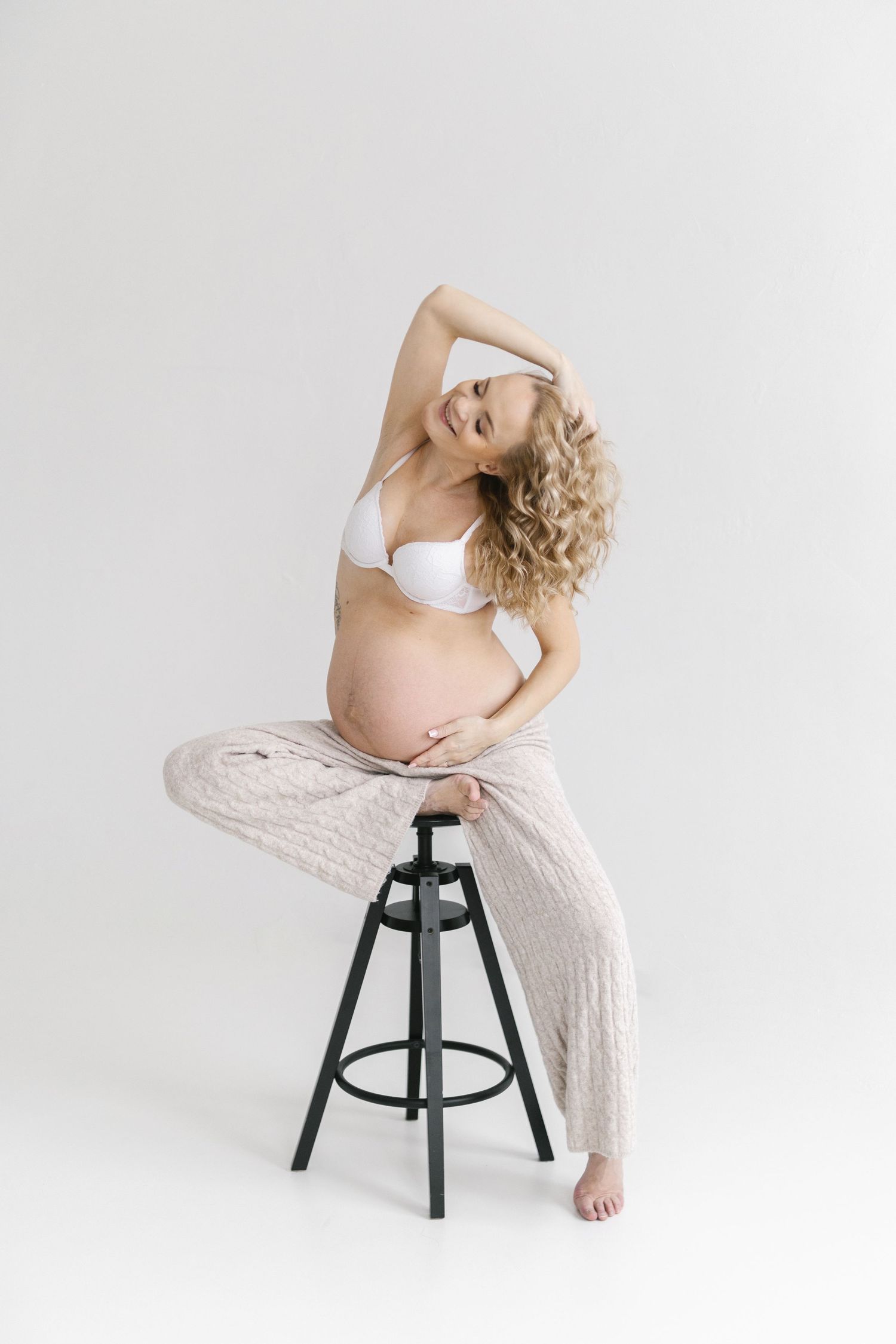 Maternity photo of an expectant mother in white bra and knit pants posing gracefully on a stool.