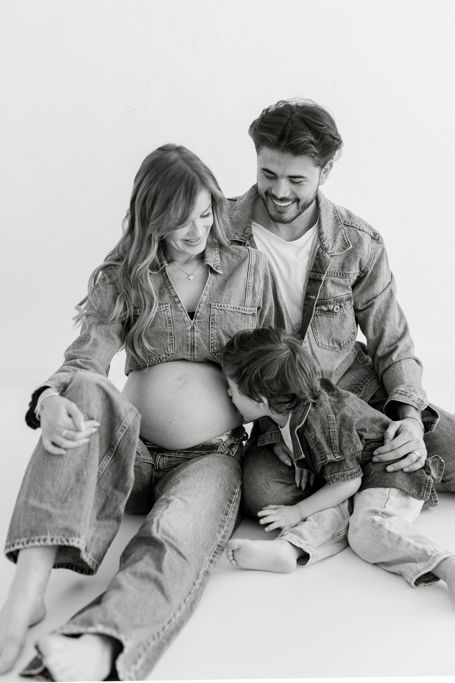 Couple in denim clothing sitting together during maternity photoshoot against white background.