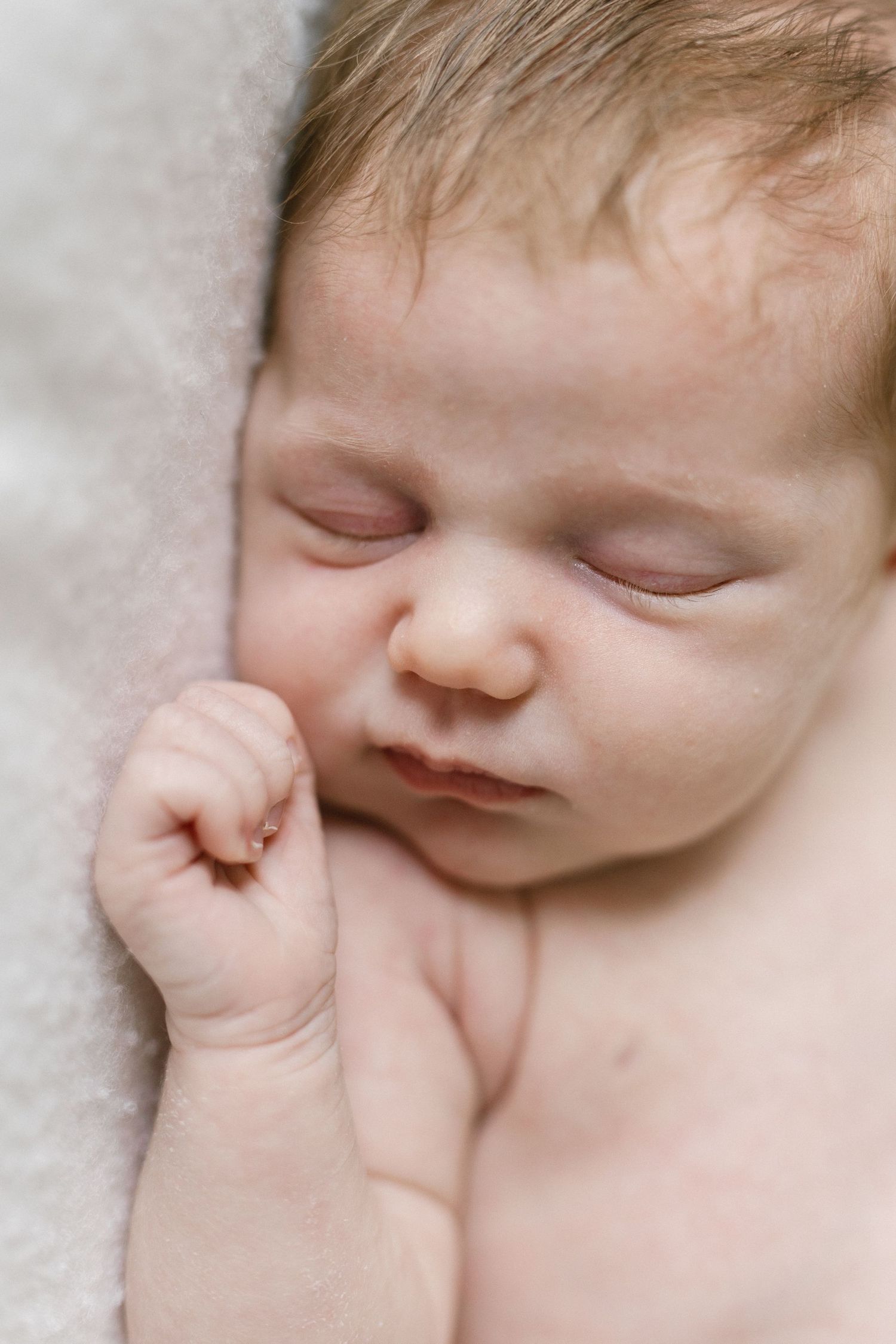 Close-up of a sleeping newborn baby with a tiny hand curled near its cheek, resting peacefully on a soft cream blanket.