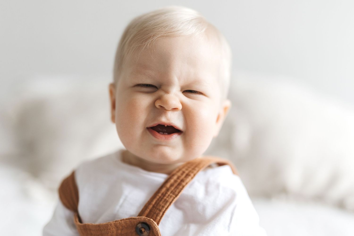 Adorable baby with blonde hair smiling and squinting, wearing brown overalls and a white shirt.