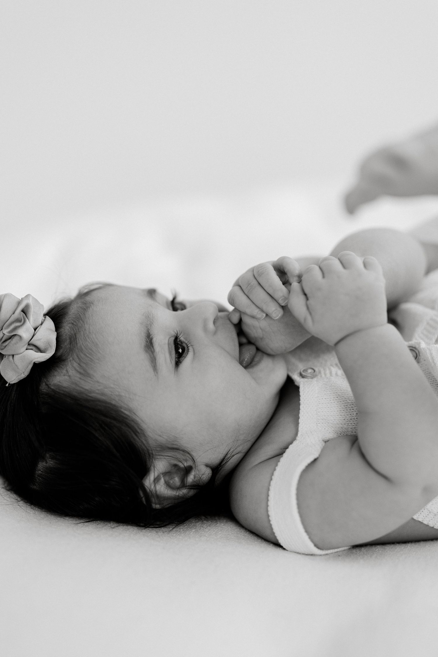 Black and white photo of an adorable baby girl lying on her back, sucking her fingers, with a flower hair clip.