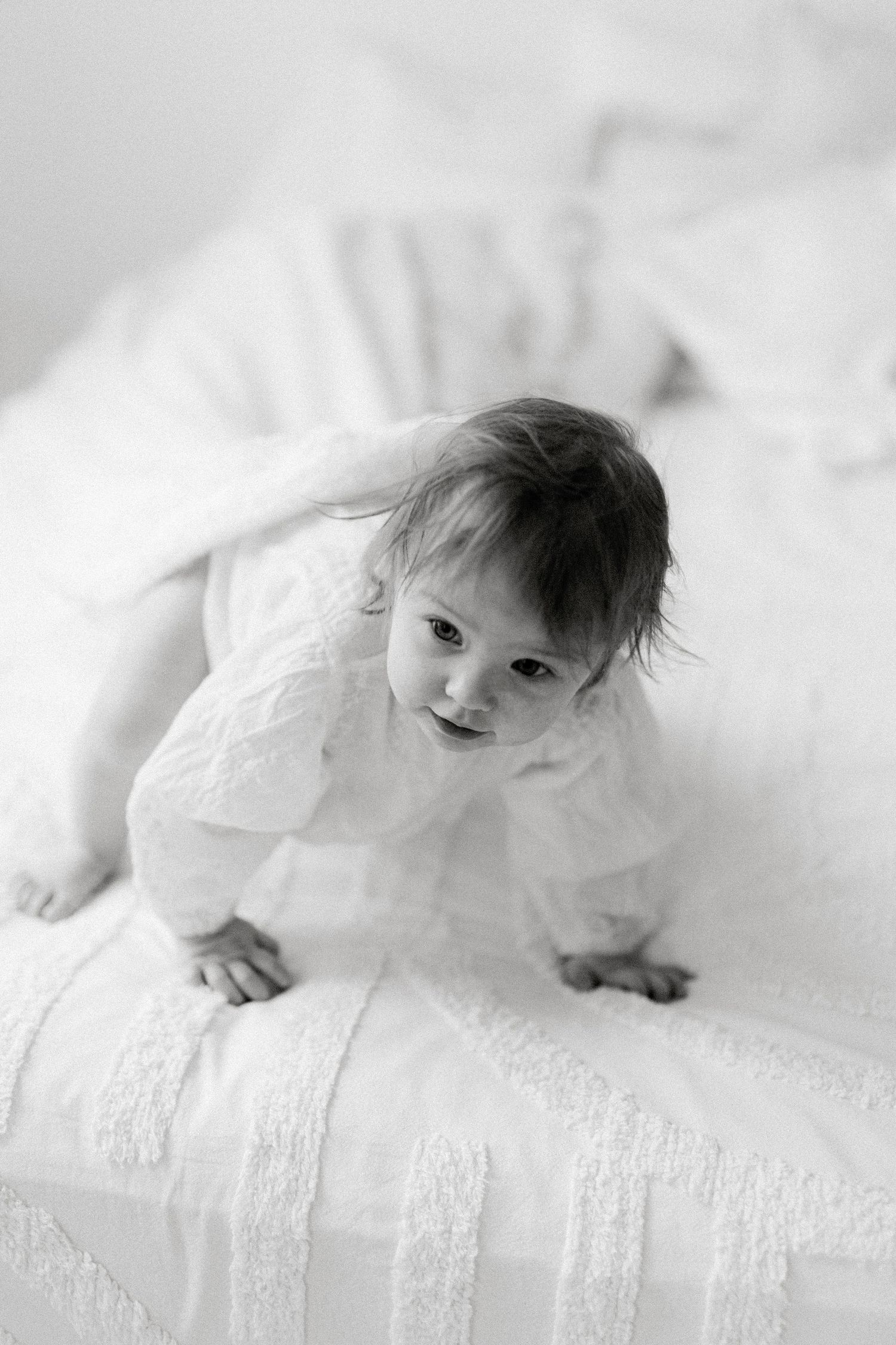 Black and white photo of a baby girl in a white lace dress crawling on a white bed.